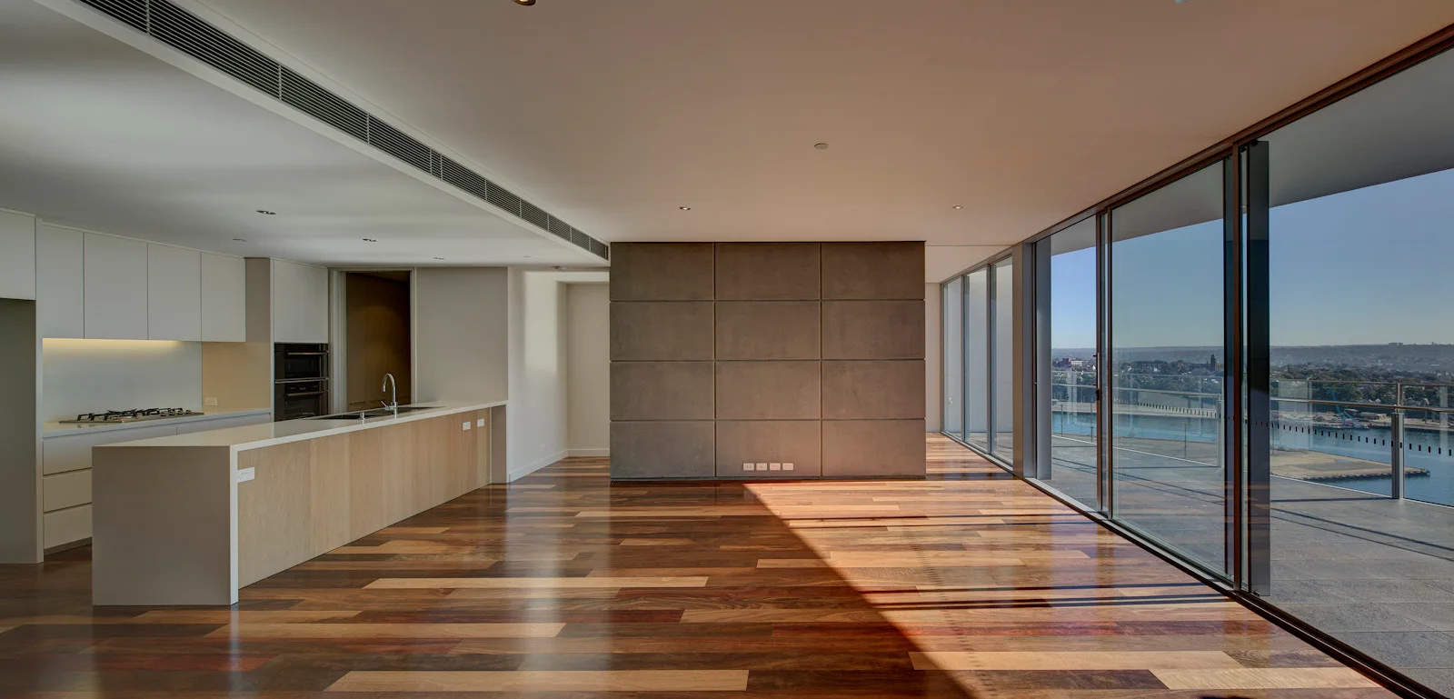 An empty open-concept kitchen features multi-toned hardwood floors, a minimalist island, a grey grid accent wall, and floor-to-ceiling glass windows.