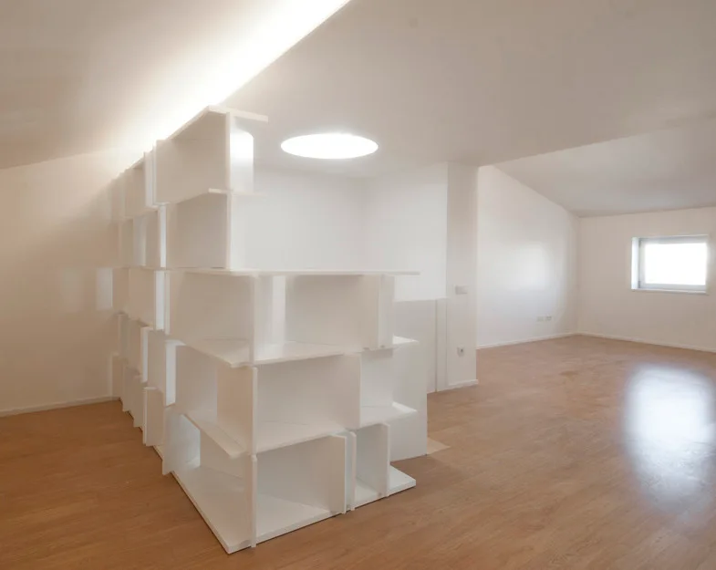 A minimalist white attic room features a geometric modular shelving unit used as a space divider, light wood flooring, and a circular skylight.