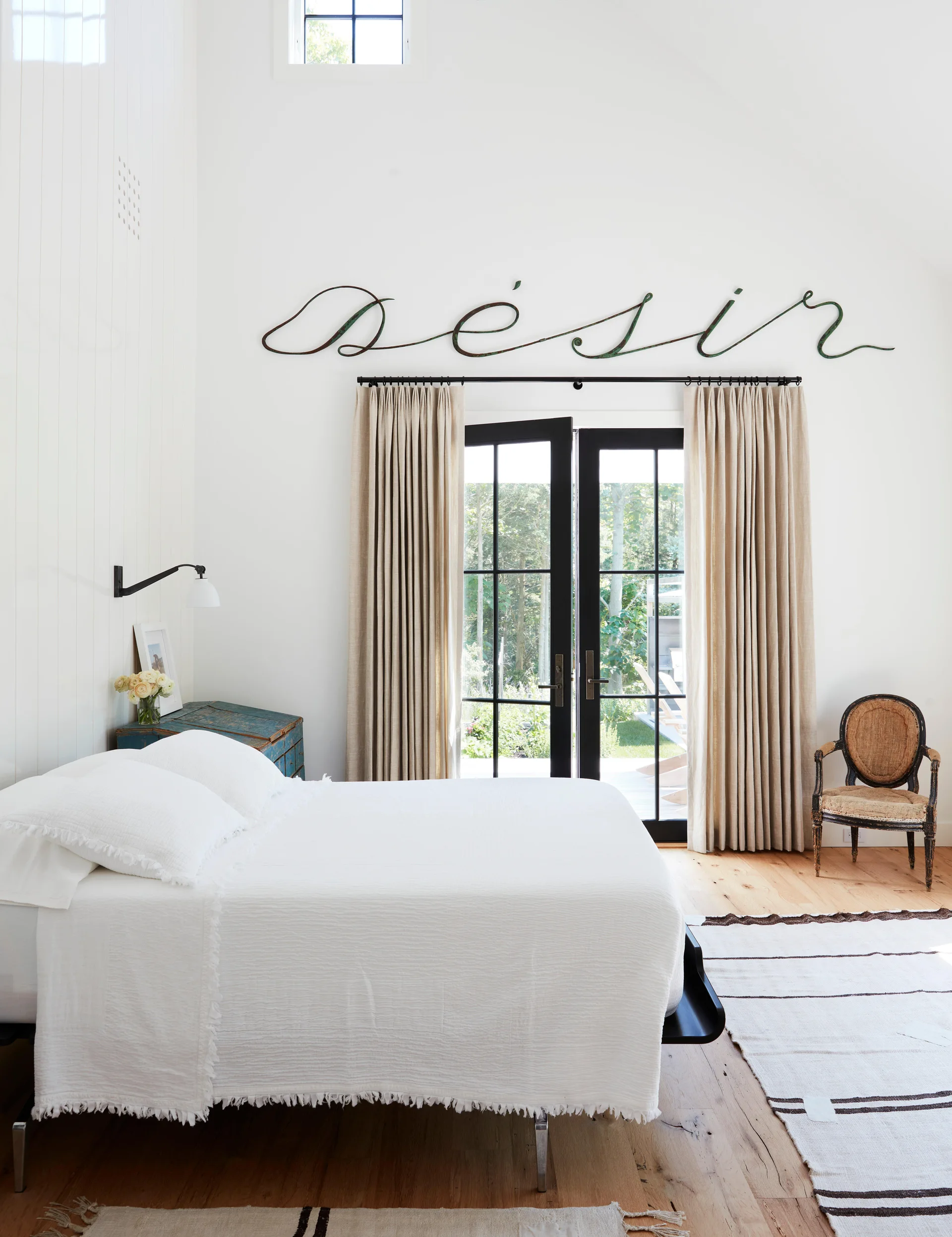 A bright bedroom featuring a white bed, vertical wood wall paneling, black-framed French doors with beige curtains, and cursive metal wall art above.
