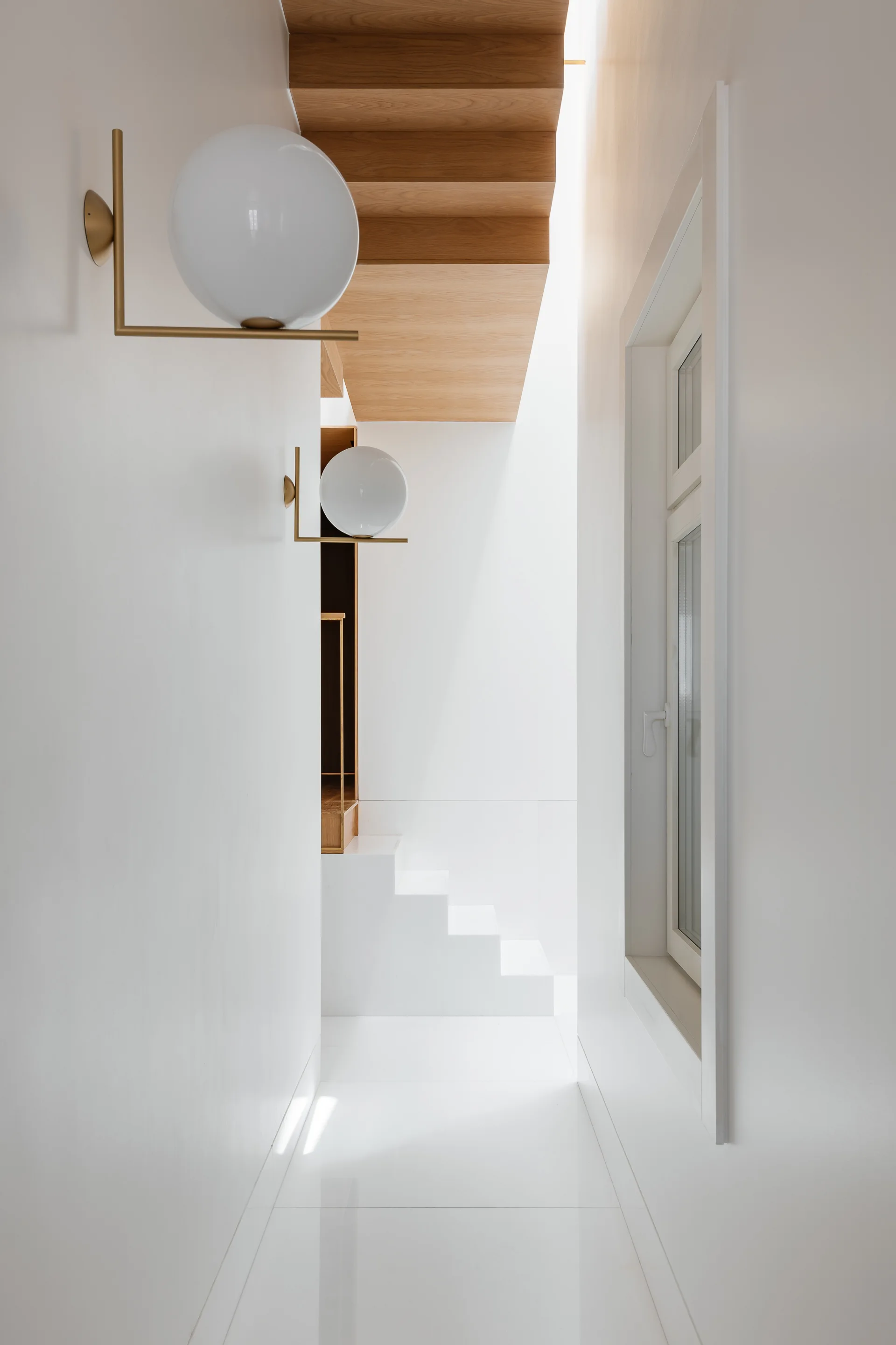 A bright white minimalist hallway features a modern staircase with wooden treads, illuminated by round brass wall sconces near a window.
