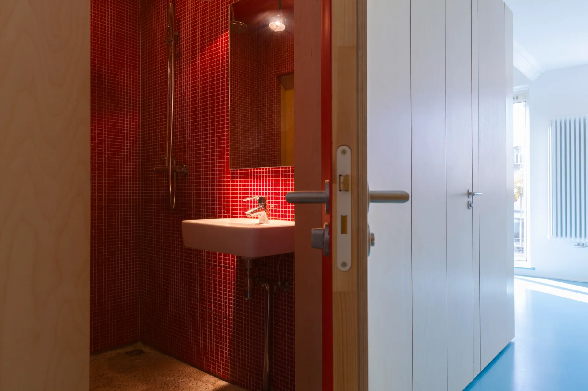 A partially open light wood door reveals a compact bathroom lined with bright red mosaic wall tiles and a small white wall-mounted sink.