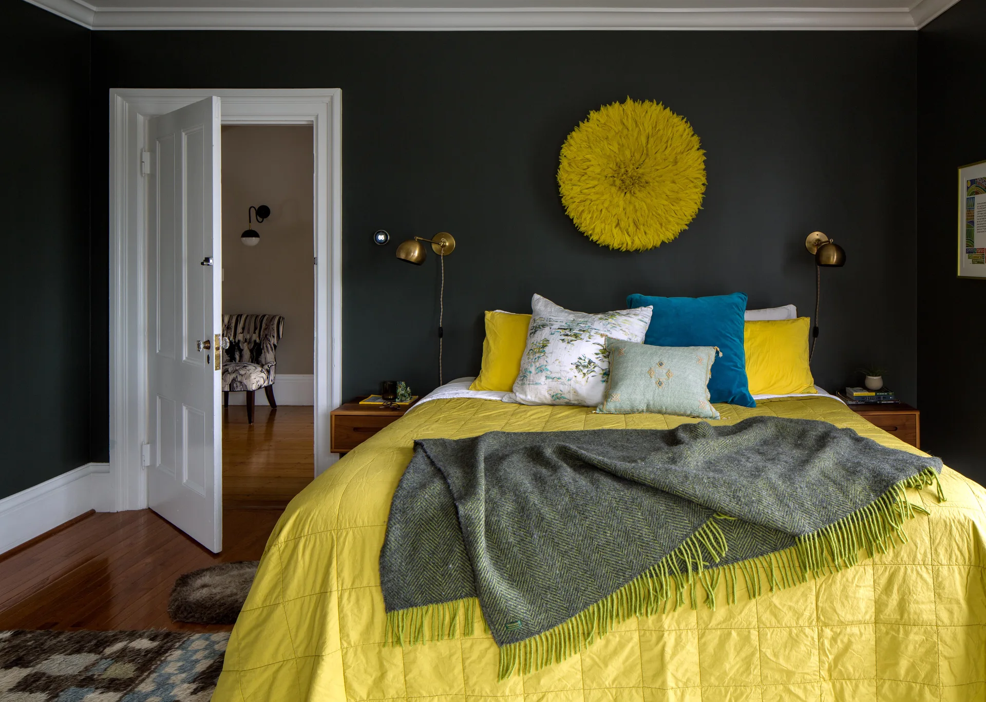 A bed with yellow bedding and colorful pillows is set against a dark charcoal wall featuring a round yellow feather decoration and brass sconces.