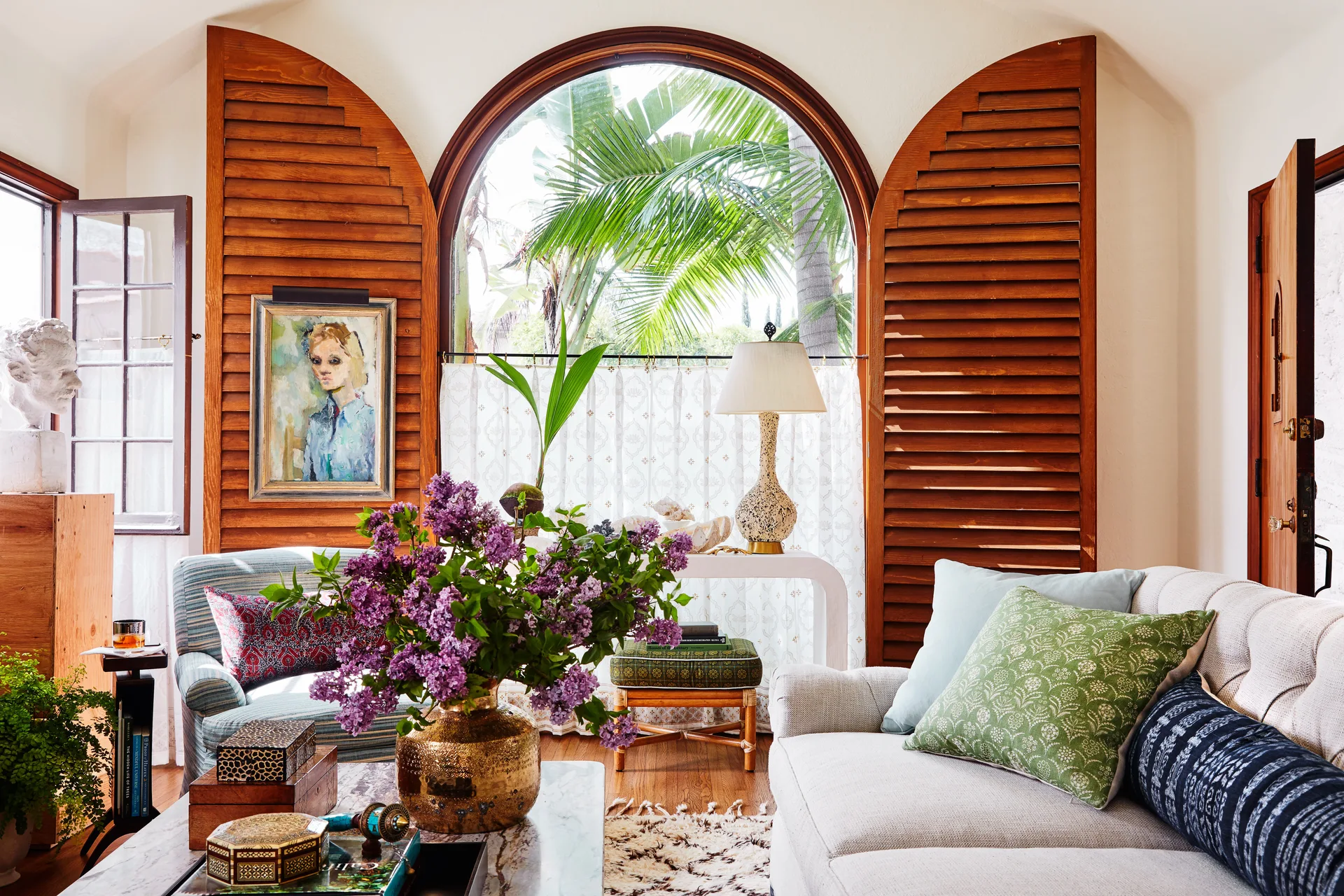 An eclectic living room features a large arched window with wooden shutters, a neutral sofa with patterned pillows, and a marble coffee table.
