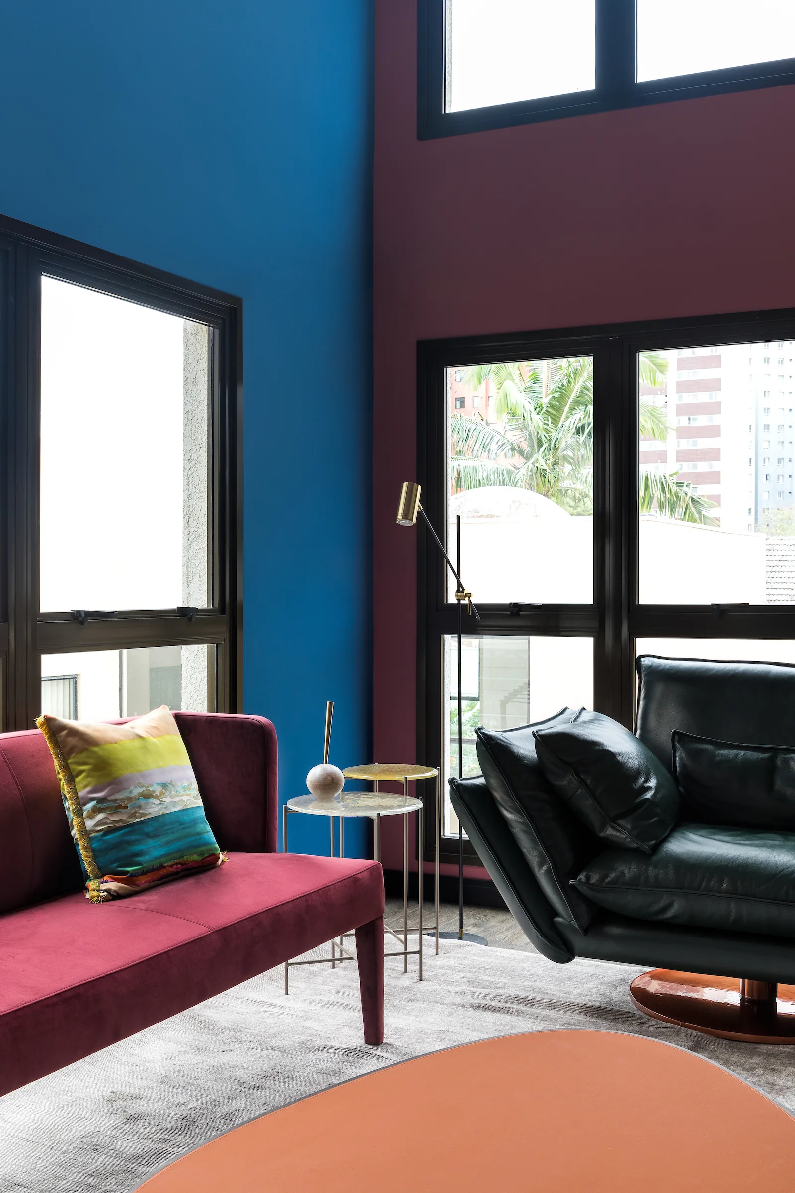 A fuchsia velvet sofa and a black leather armchair sit on a grey rug against blue and plum color-blocked walls near a tall window.