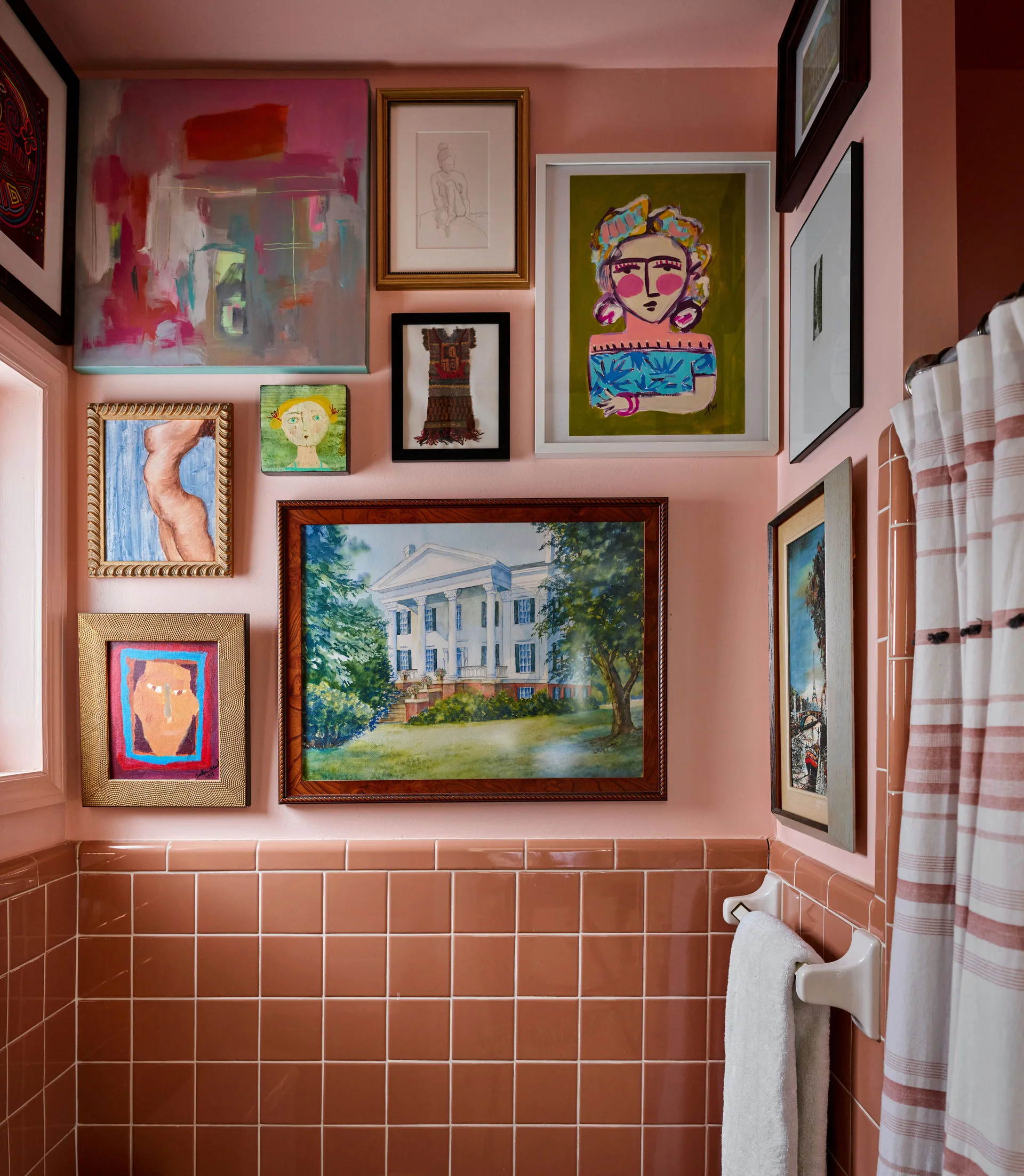 A bathroom features glossy pink wall tiles on the bottom half and an eclectic collection of framed art on the pink painted wall above.