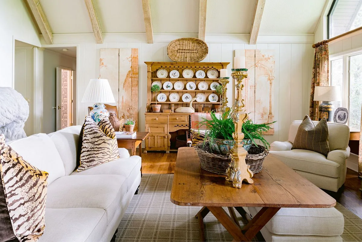 A cozy cottage living room with a cream sofa, a wooden hutch displaying plates, and a vaulted ceiling with exposed beams.