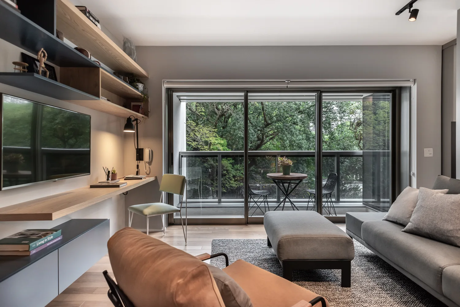 A contemporary living room features a gray sofa, a floating wood desk with built-in shelves, and glass sliding doors opening to a lush balcony.