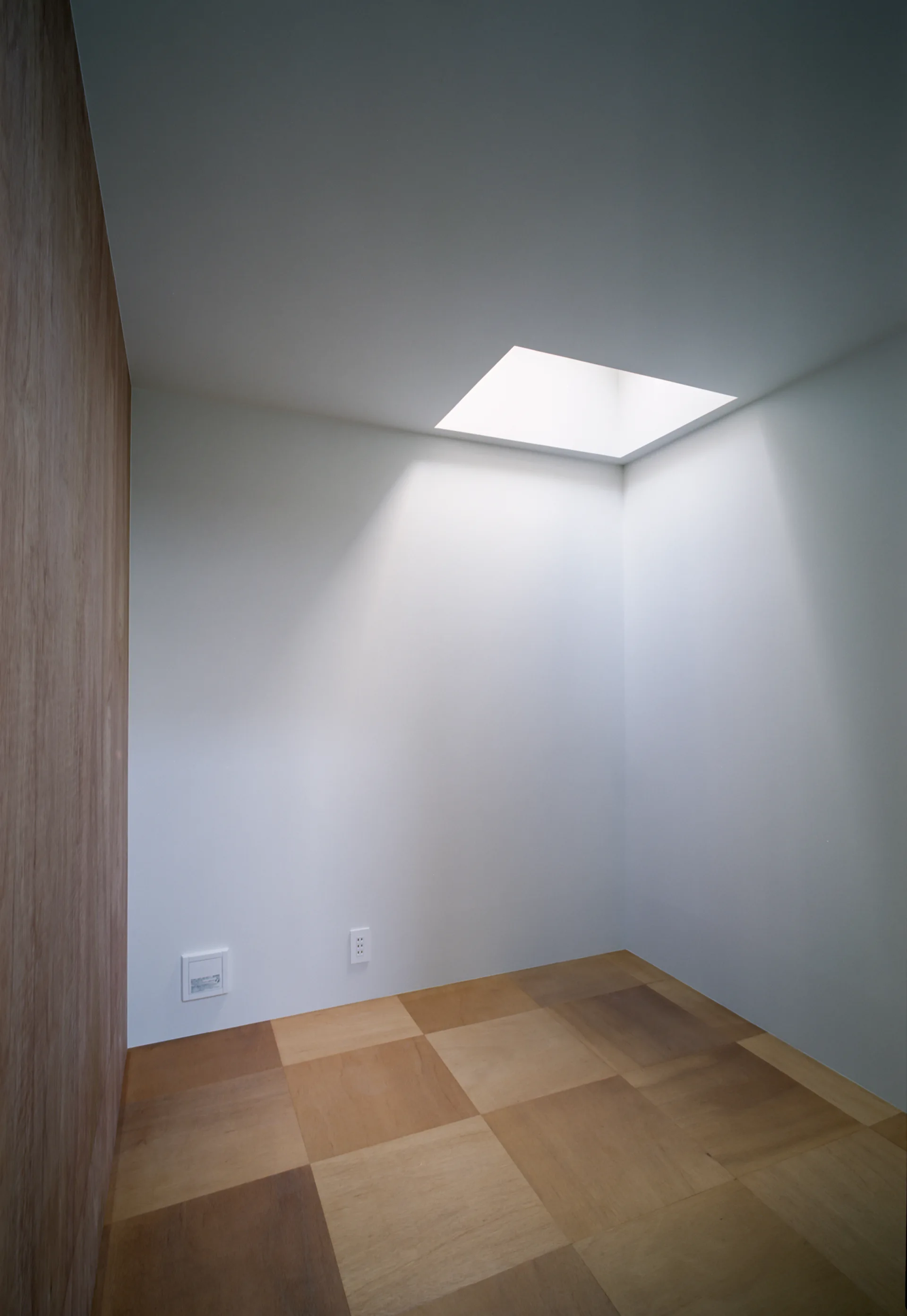 A minimalist empty room corner features white walls, a light wood paneled wall, and checkerboard patterned wood floor tiles below a ceiling skylight.