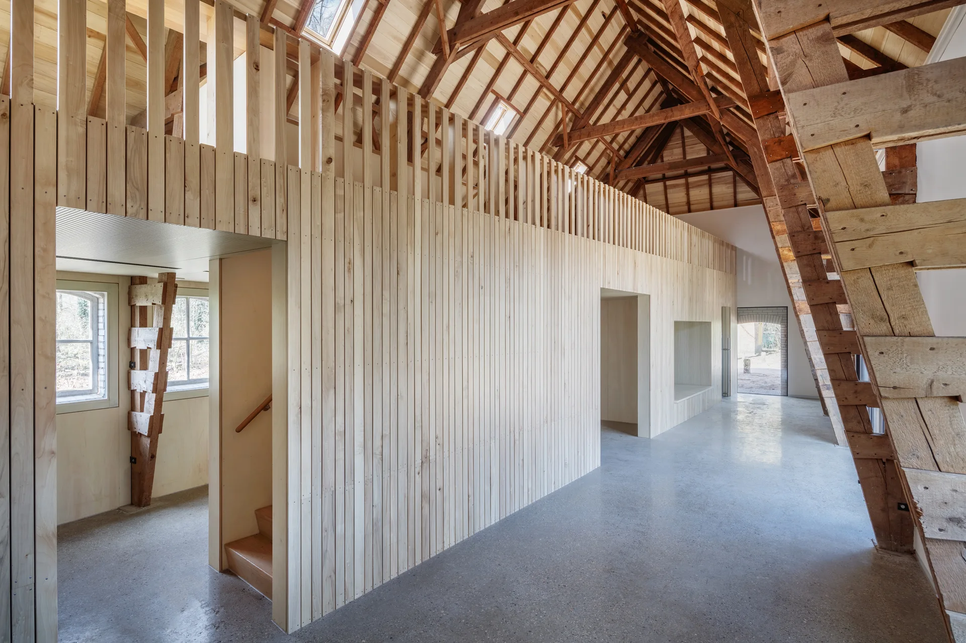 A modern barn interior features a light wood vertical slatted partition wall beneath an exposed timber truss ceiling on a polished concrete floor.