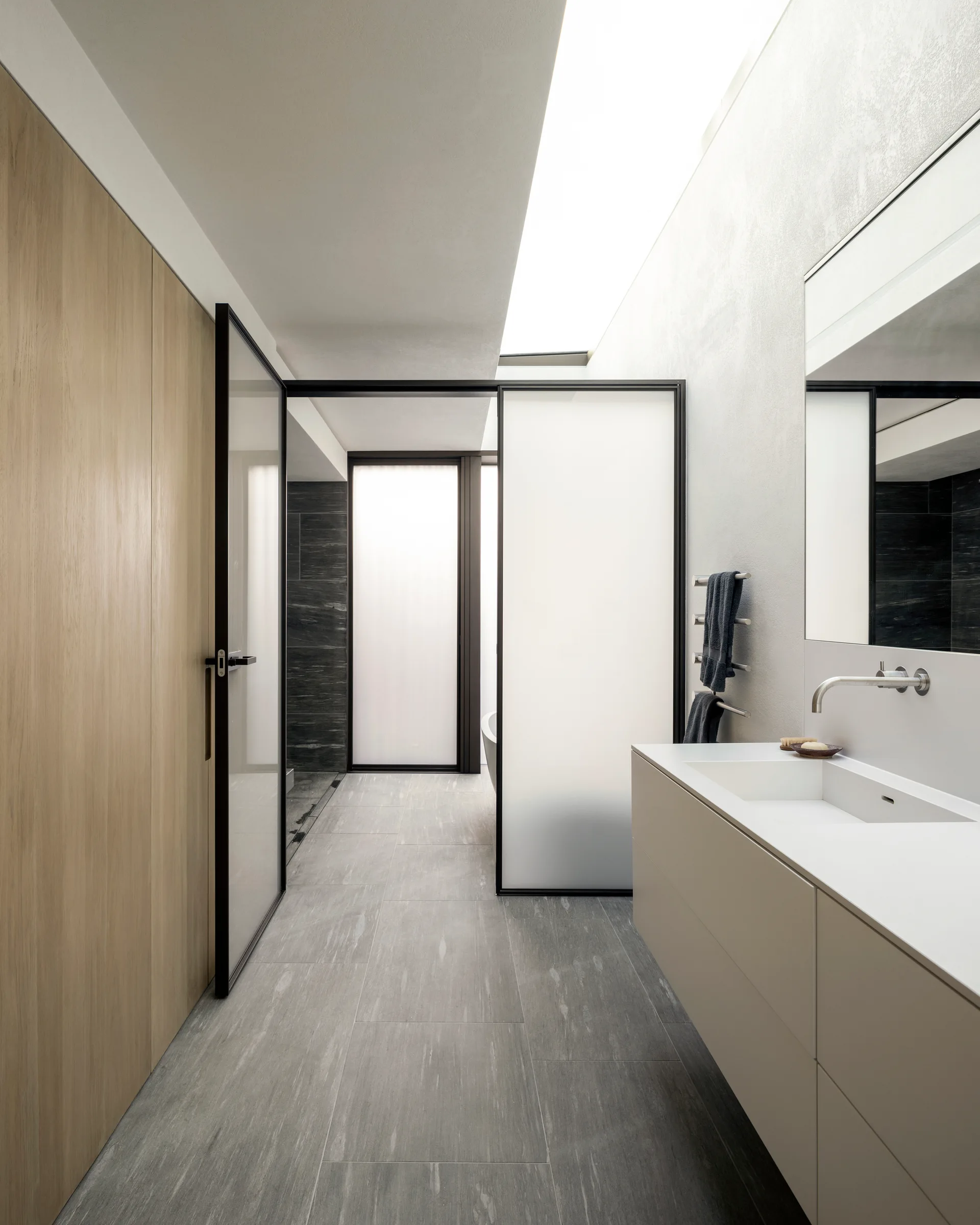 A minimalist bathroom features a sleek wall-mounted vanity, gray floor tiles, a wood-paneled wall, and a frosted glass partition beneath a bright skylight.