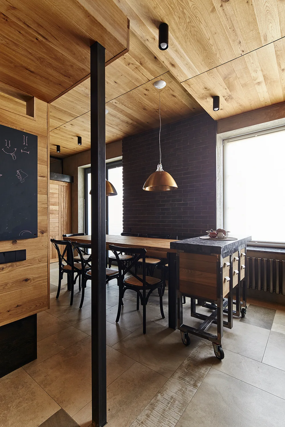 An industrial dining area with a long wooden table, black crossback chairs, a dark brick accent wall, and a wood plank ceiling.
