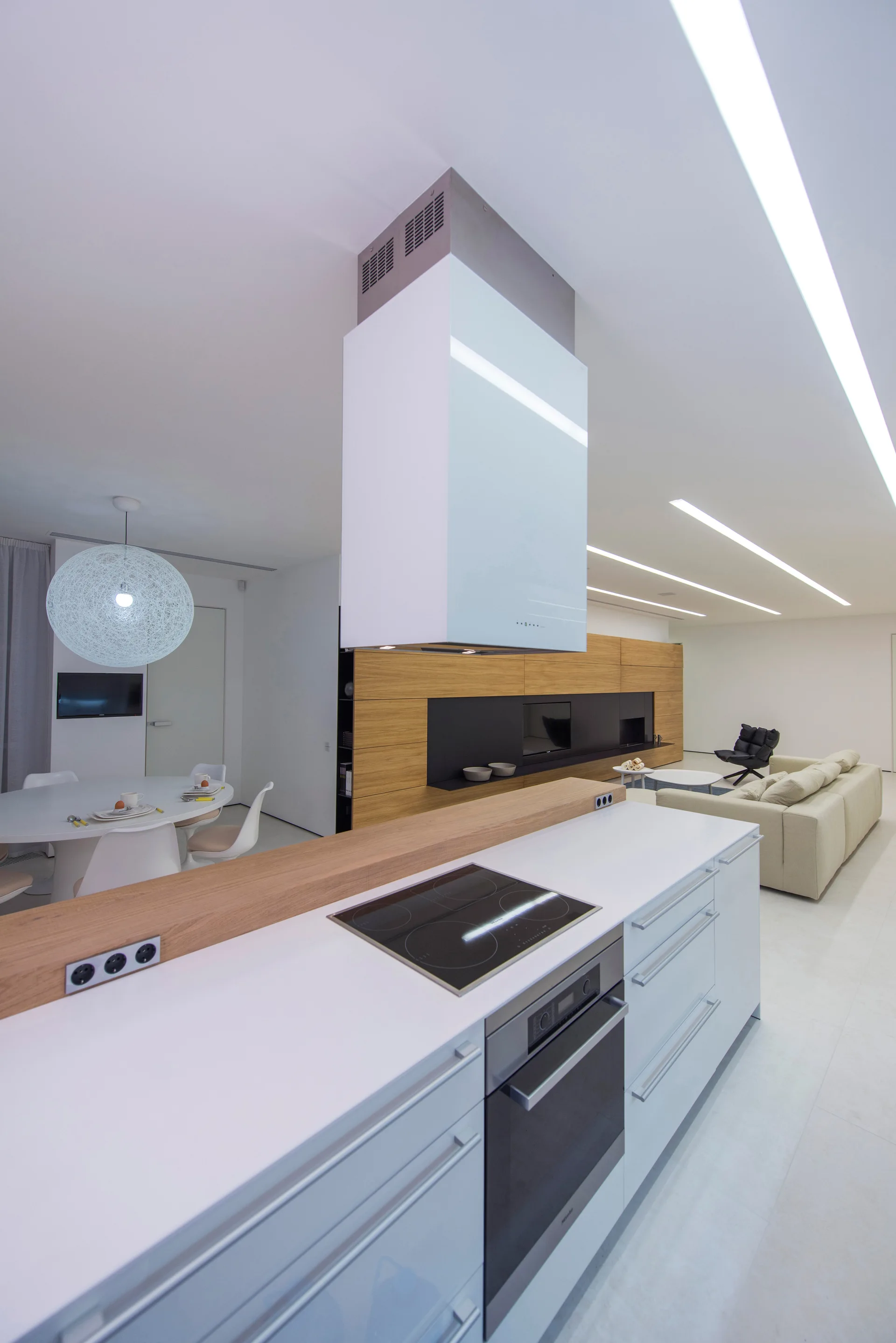 A modern open-concept kitchen featuring a white island with a cooktop, a suspended range hood, linear ceiling lights, and wood-paneled wall storage.