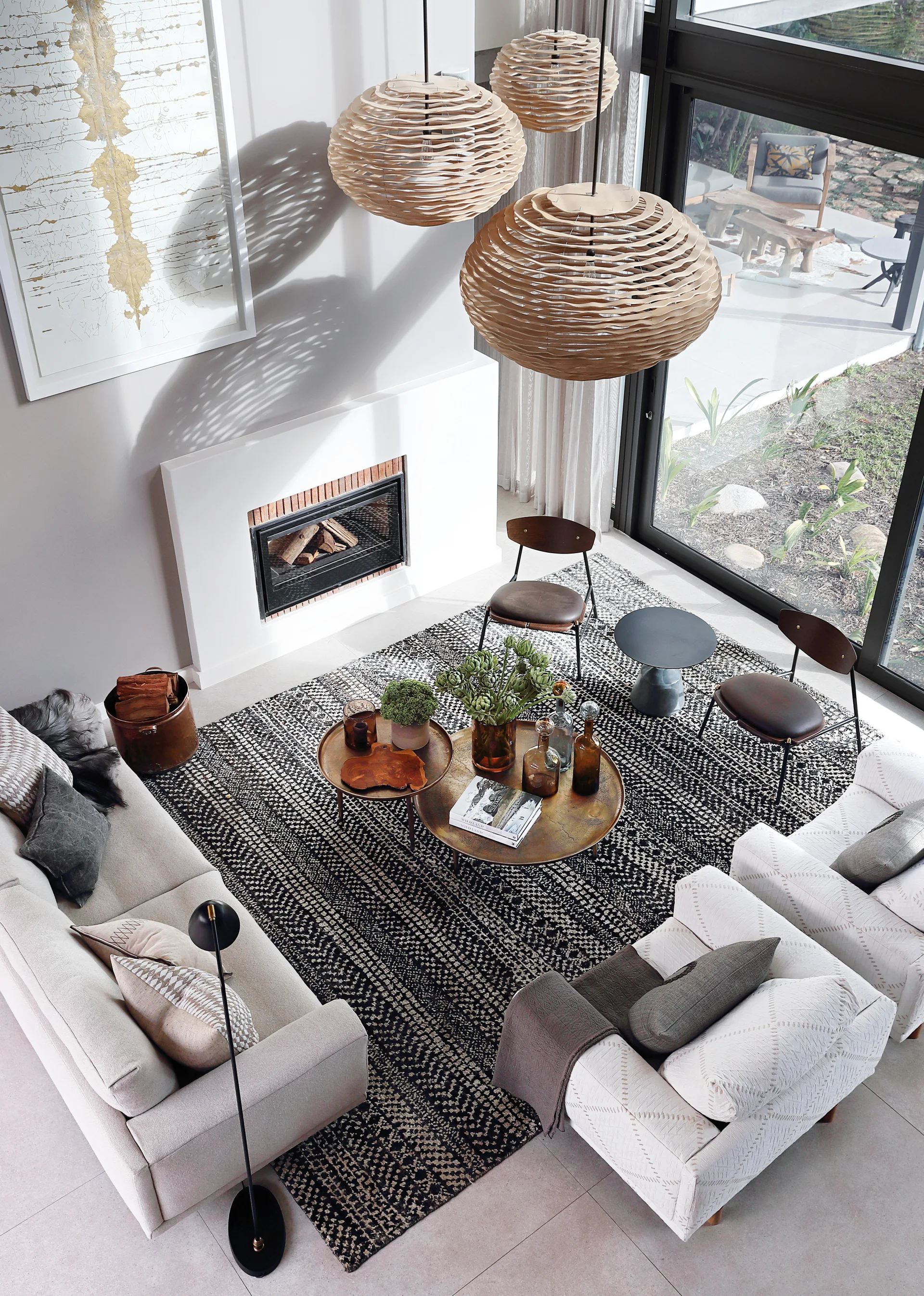 A high-angle view of a modern living room featuring three woven wood pendant lights, a black and white patterned rug, and a minimalist white fireplace.