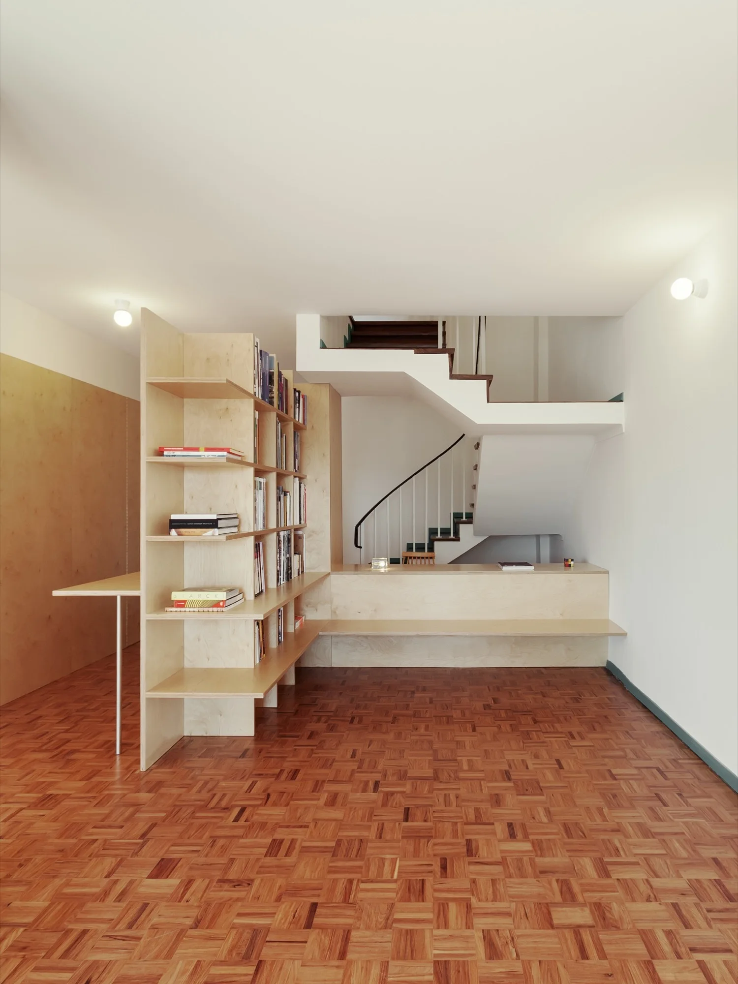 An L-shaped custom plywood bookshelf and low bench stand on a parquet wood floor near a modern white staircase.