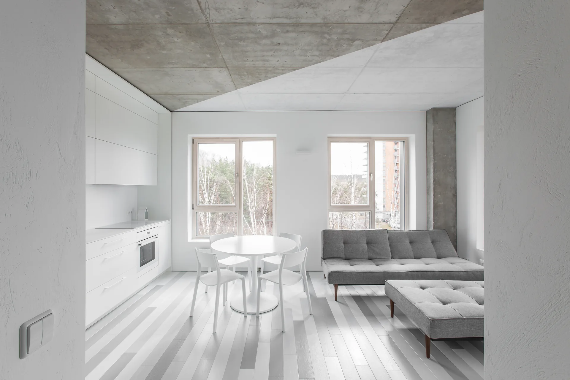 An open-plan living room and kitchen features white minimalist cabinetry, a round dining table, a grey sofa, and a striped wood floor.