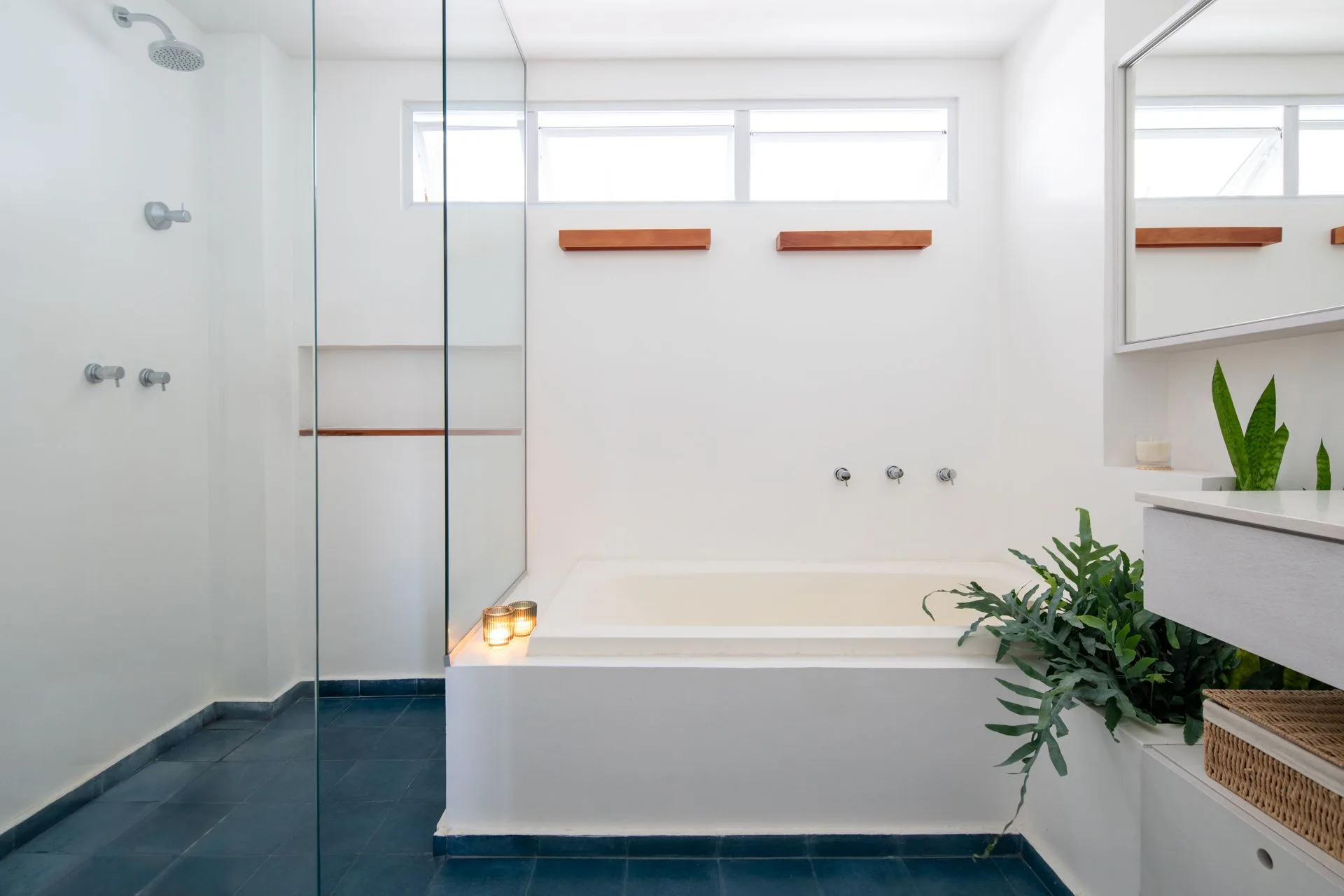A minimalist white bathroom featuring a built-in tub, a glass-enclosed shower, dark blue tiled flooring, and high clerestory windows.