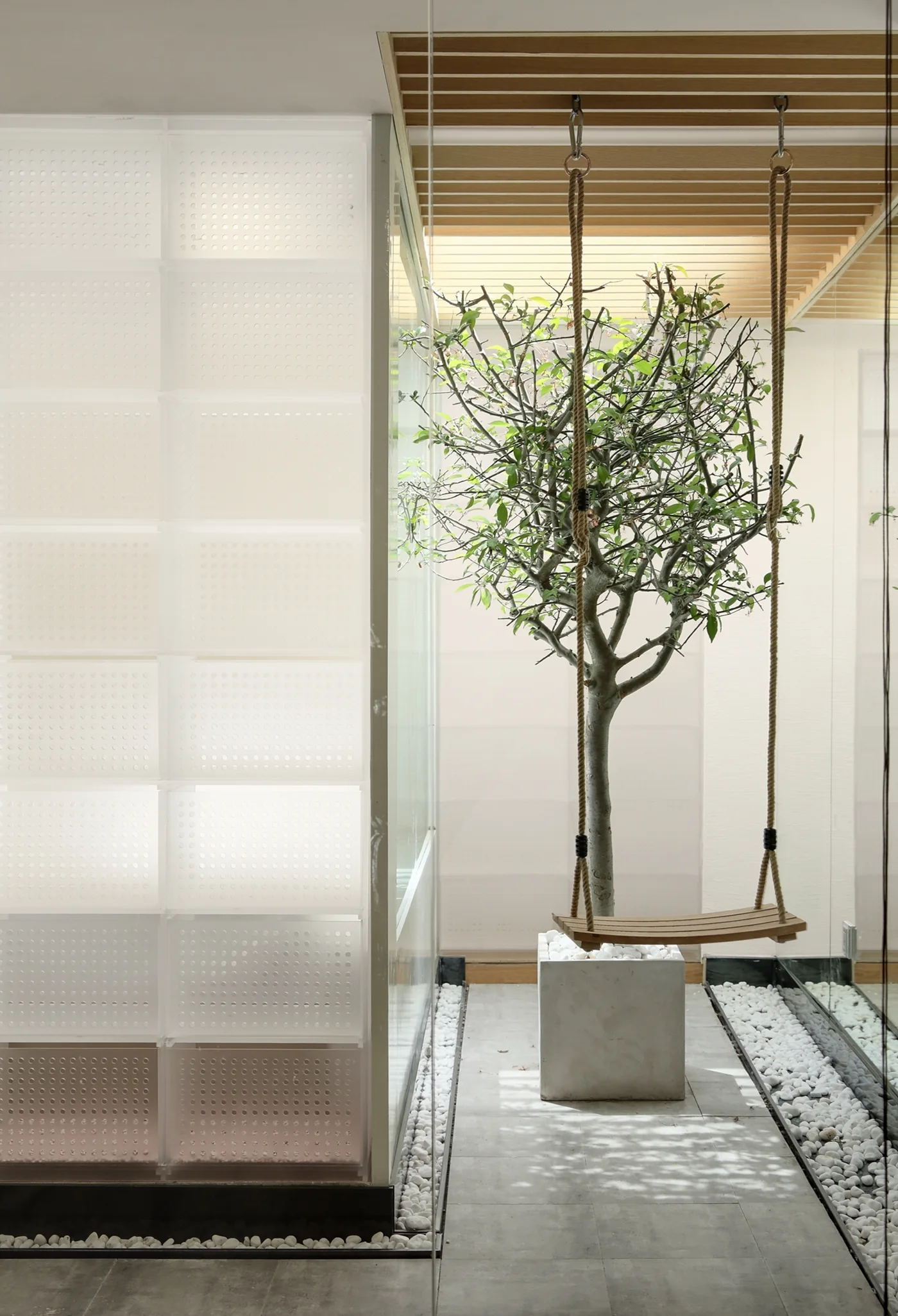 A potted tree with a hanging wooden swing sits in a minimalist indoor courtyard under a slatted ceiling beside a white perforated wall.