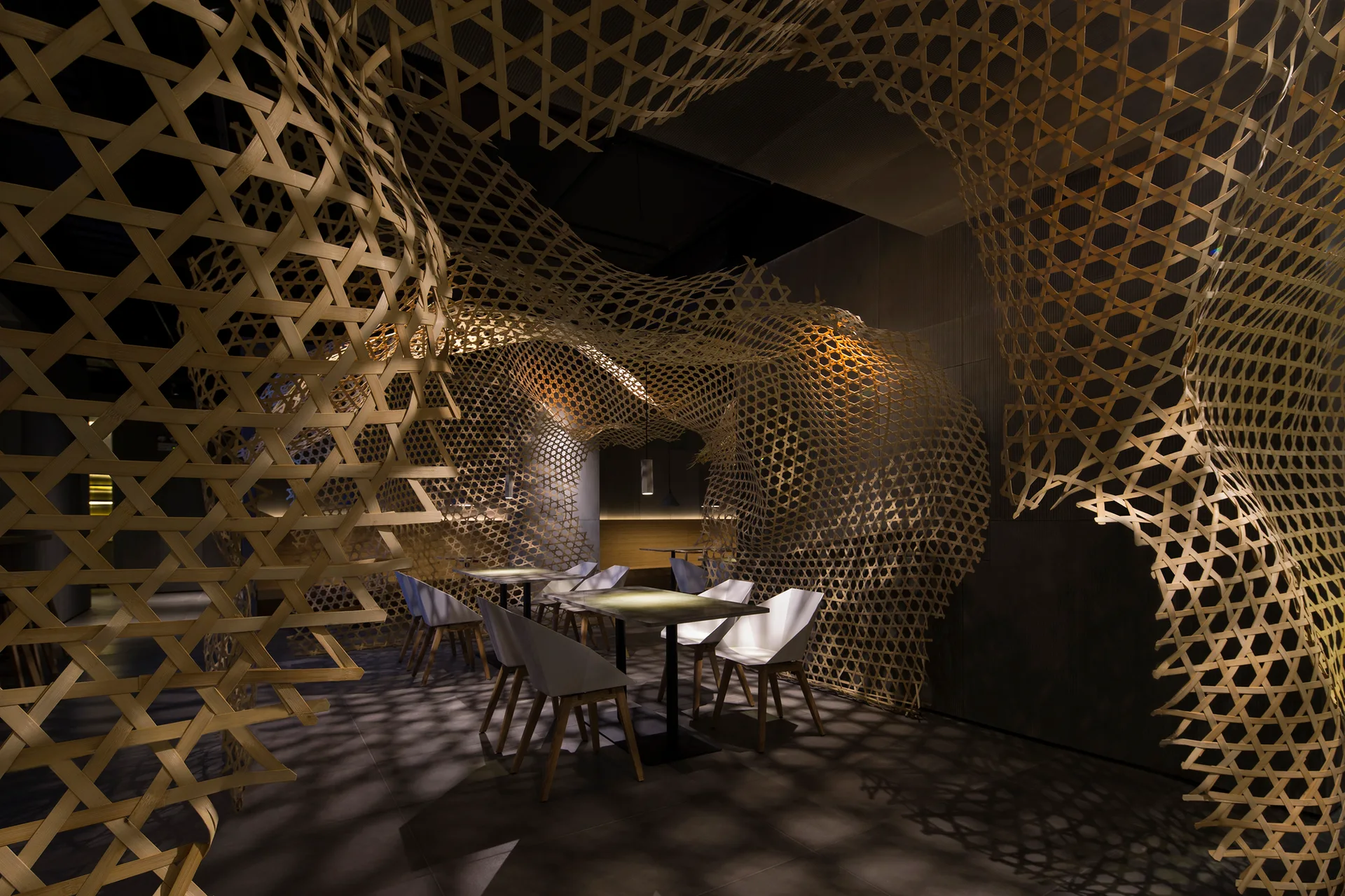 A modern dining area featuring white geometric chairs, square tables, and a dramatic, woven bamboo latticework installation that spans the ceiling and walls.