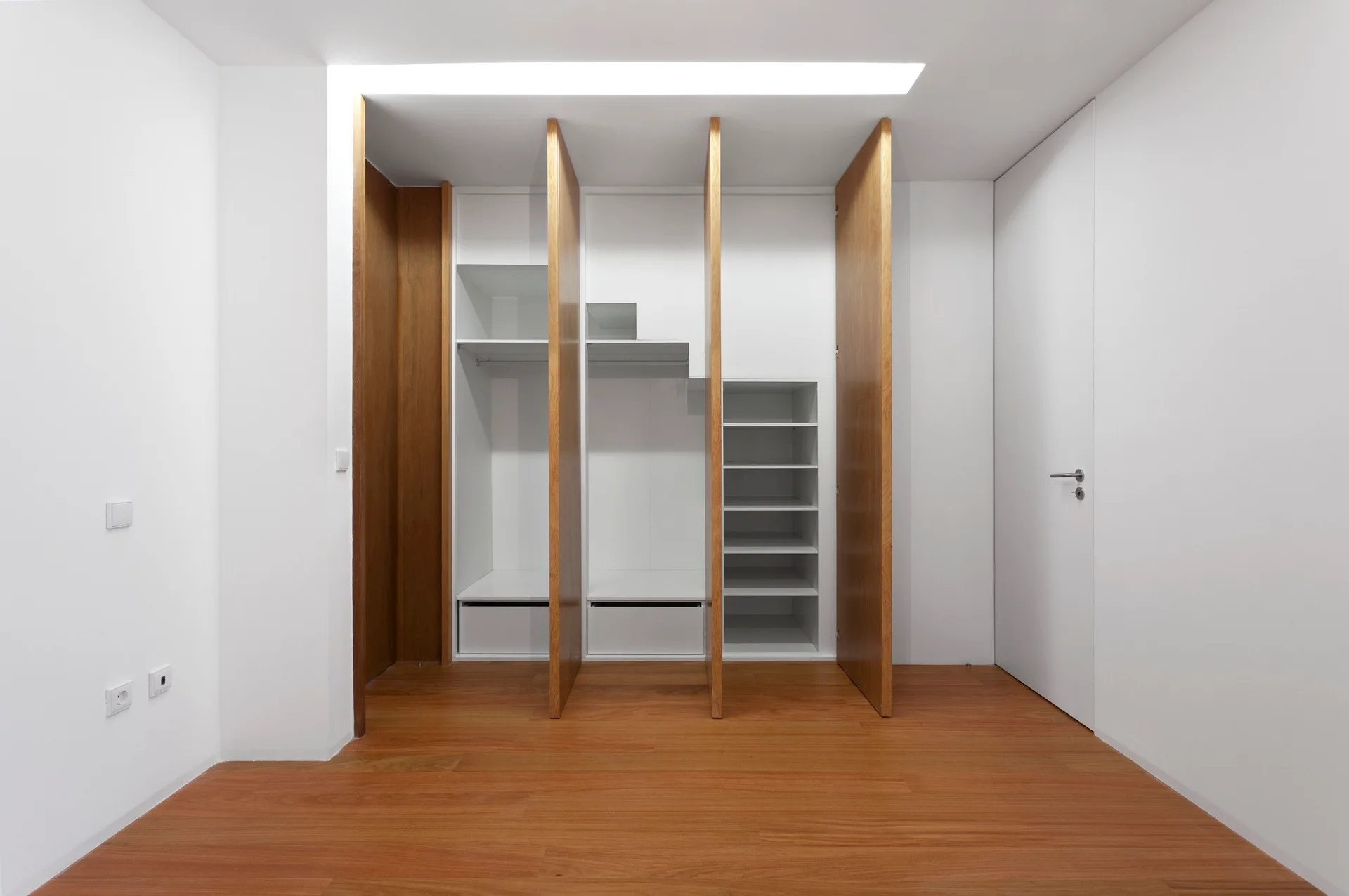 A built-in wardrobe features white shelving units separated by vertical wood panels, set on medium-toned wood flooring beneath a recessed ceiling light.