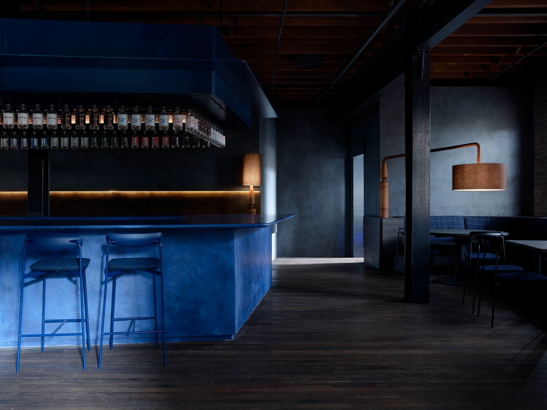 A dark industrial bar interior features a saturated blue counter with matching bar stools, textured dark grey walls, and exposed wooden ceiling beams.