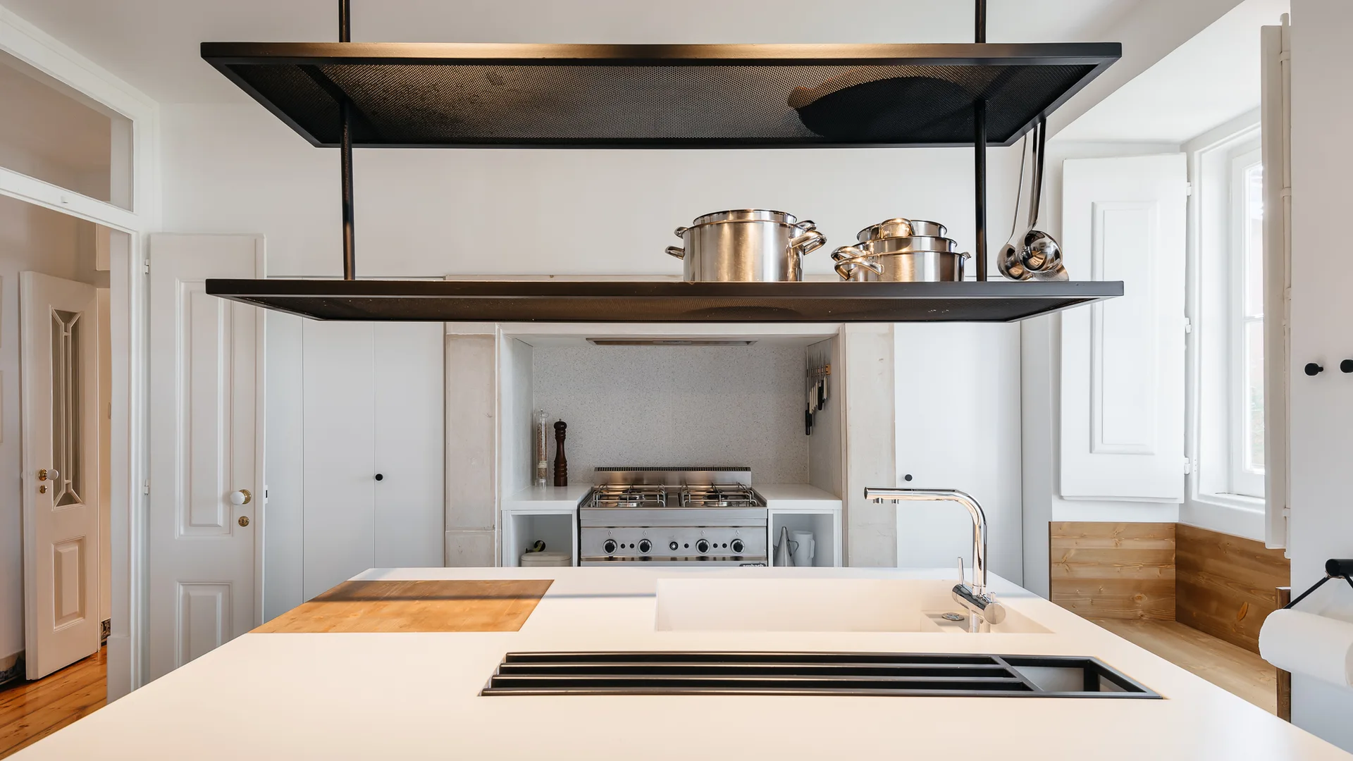 A bright white kitchen features a central island with a chrome faucet, beneath a suspended black metal rack holding stainless steel pots.