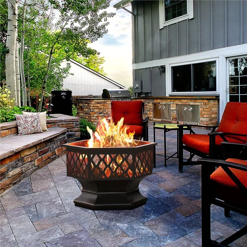 A stone paver patio features a central black hexagonal fire pit surrounded by outdoor armchairs with red cushions and a built-in stone bench.