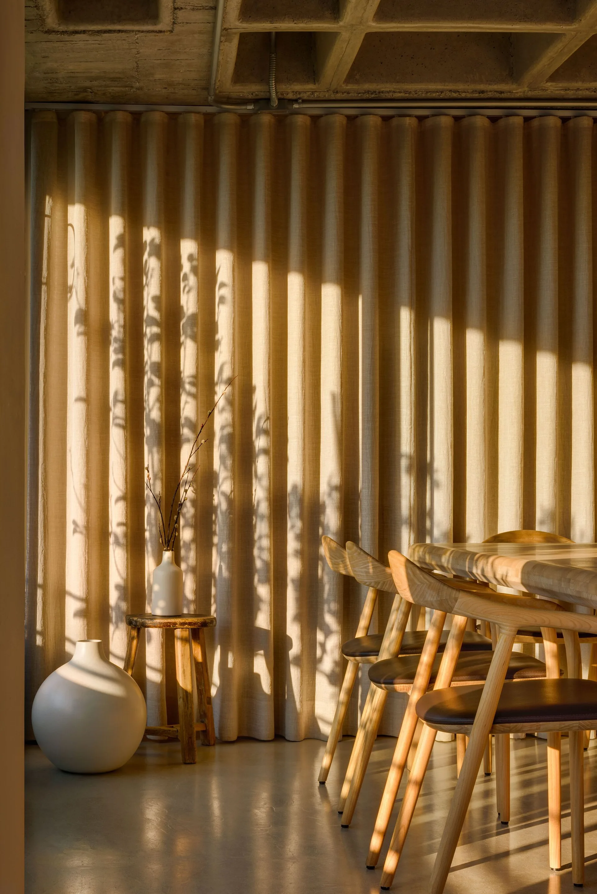 A light wood dining set sits before floor-to-ceiling beige pleated curtains under an exposed concrete waffle ceiling with dramatic branch shadows.