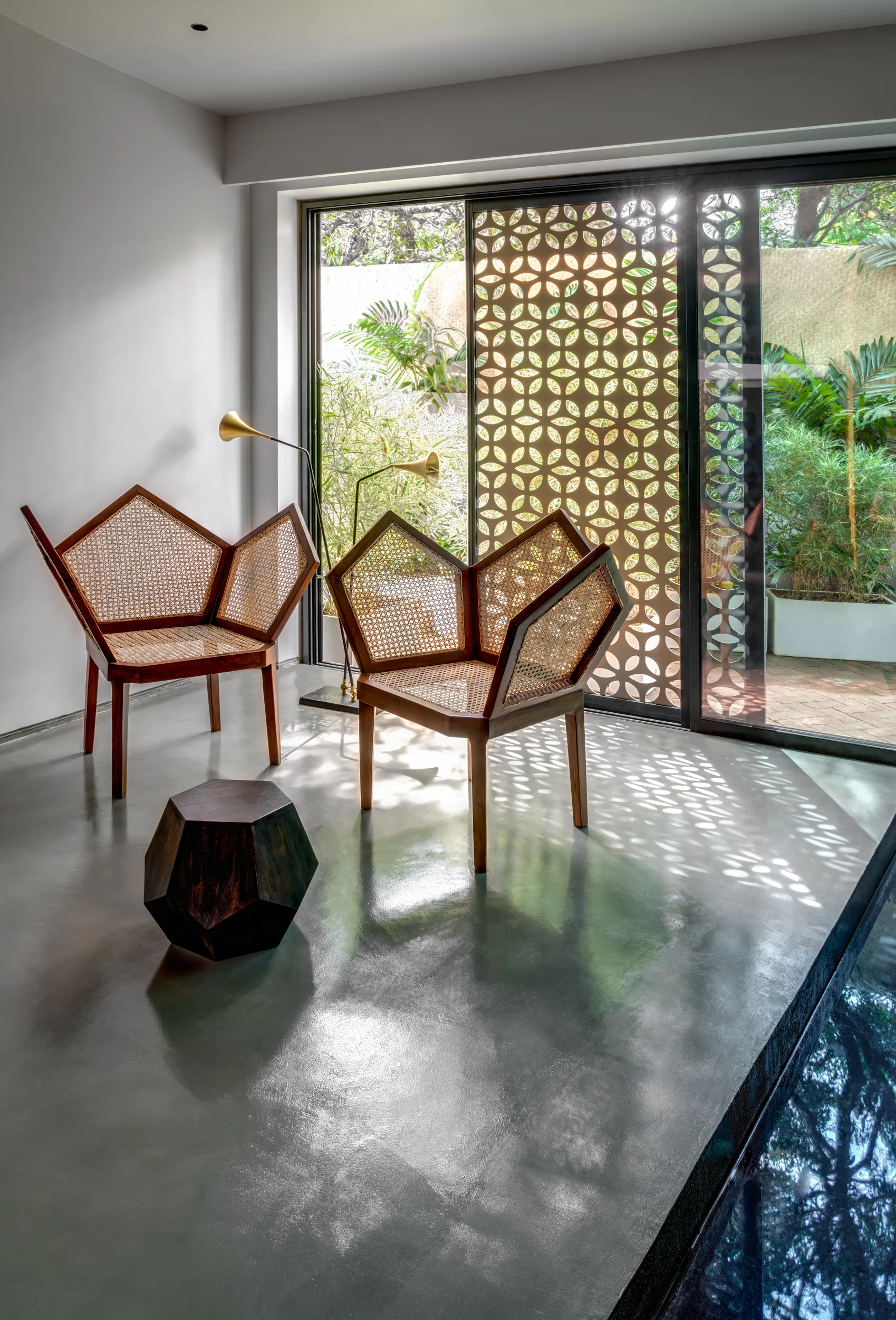 Two geometric wooden armchairs with cane backs sit on a glossy grey concrete floor next to a glass door with a patterned decorative screen.