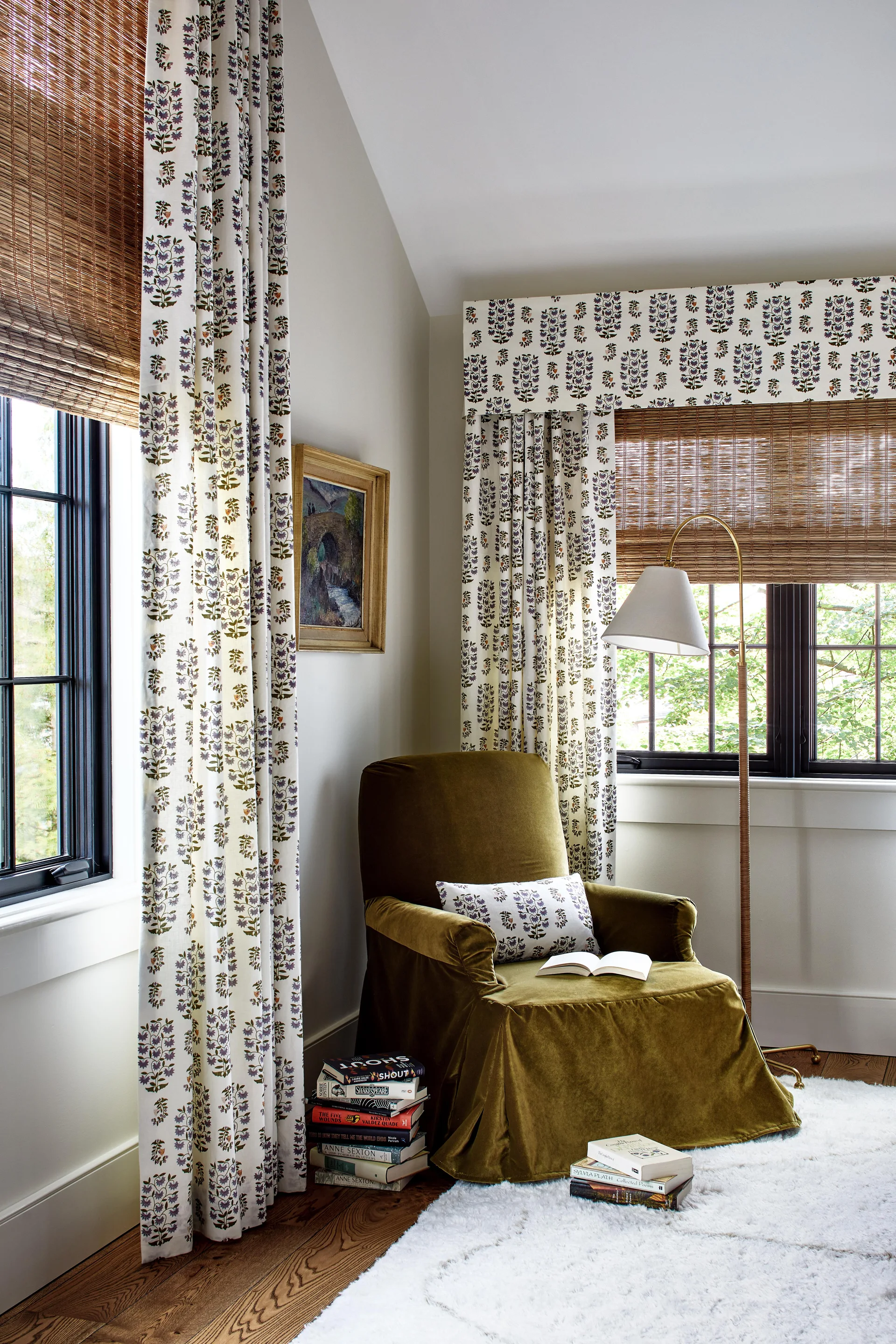 An olive green velvet armchair sits in a corner reading nook beside windows styled with botanical-print curtains and woven wood shades.