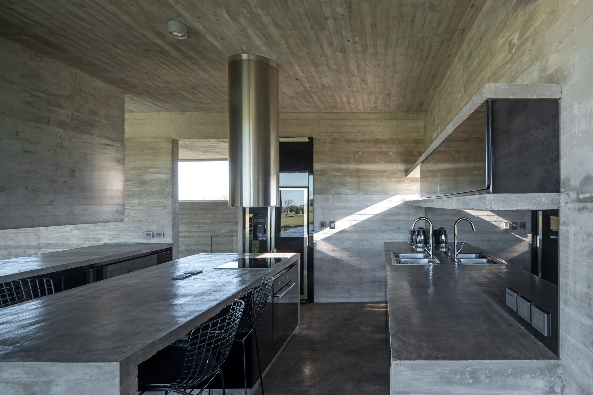 An industrial kitchen features board-formed concrete walls and ceiling, a matching concrete island with black wire chairs, and a stainless steel range hood.