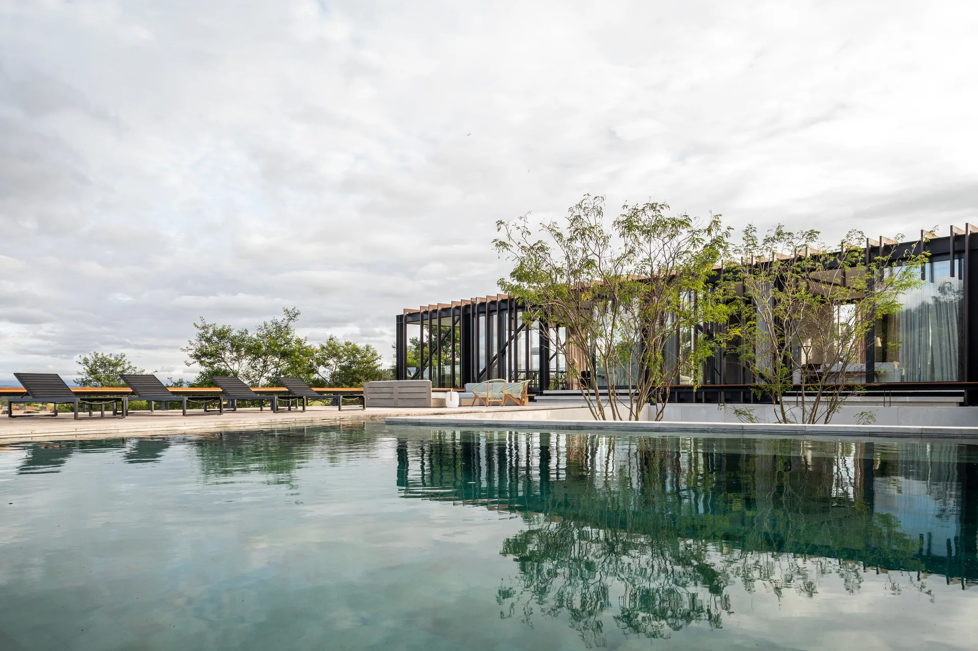 A rectangular swimming pool reflects an overcast sky next to a wooden deck lined with dark loungers and a modern glass-walled pavilion.
