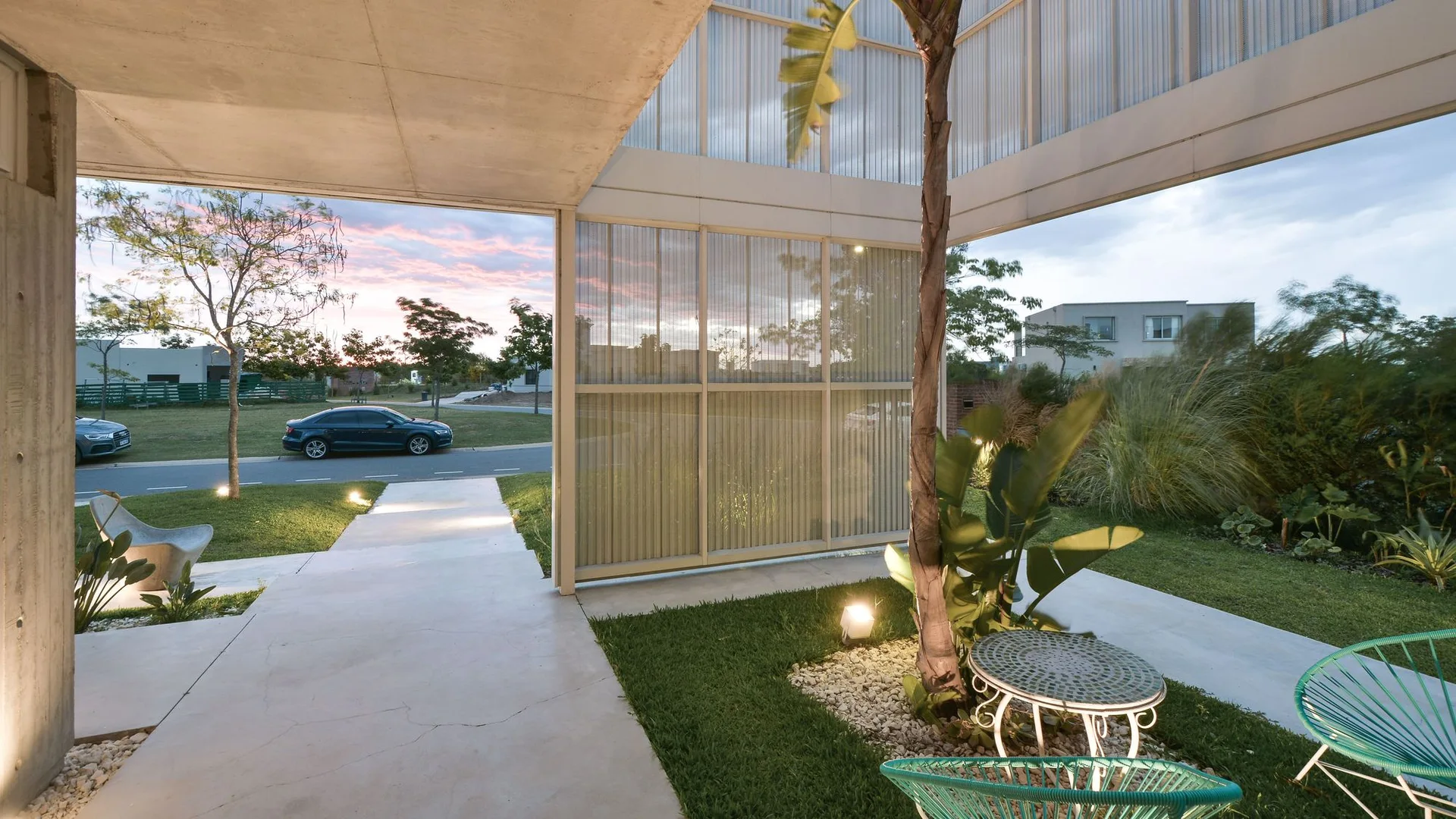 A modern covered patio with a concrete ceiling, corrugated translucent screens, green string chairs, and ground lighting in a grassy garden.