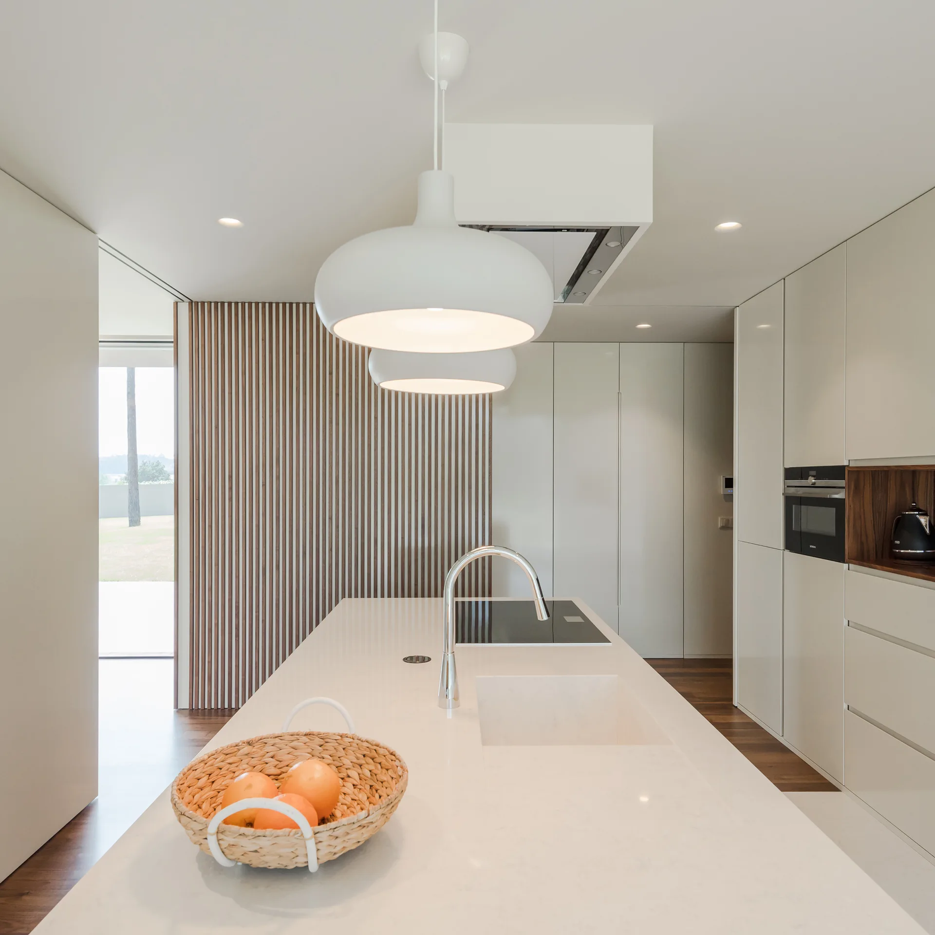 A modern kitchen features a white island with a sink, white dome pendant lights, and a warm vertical wood-slatted accent wall in the background.