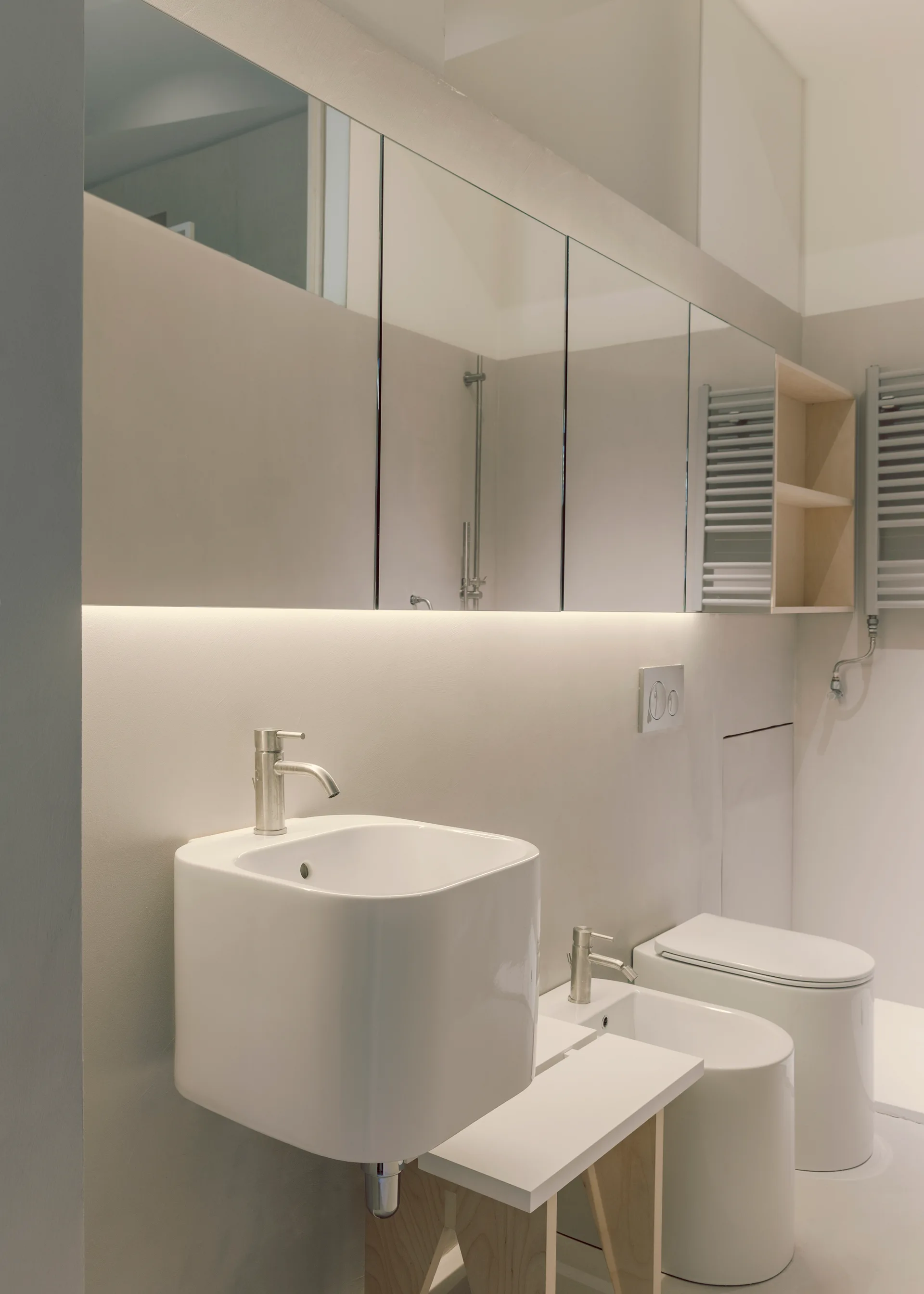 A modern grey bathroom featuring a white floating square sink, a matching bidet, and a large mirrored cabinet with integrated under-cabinet lighting.