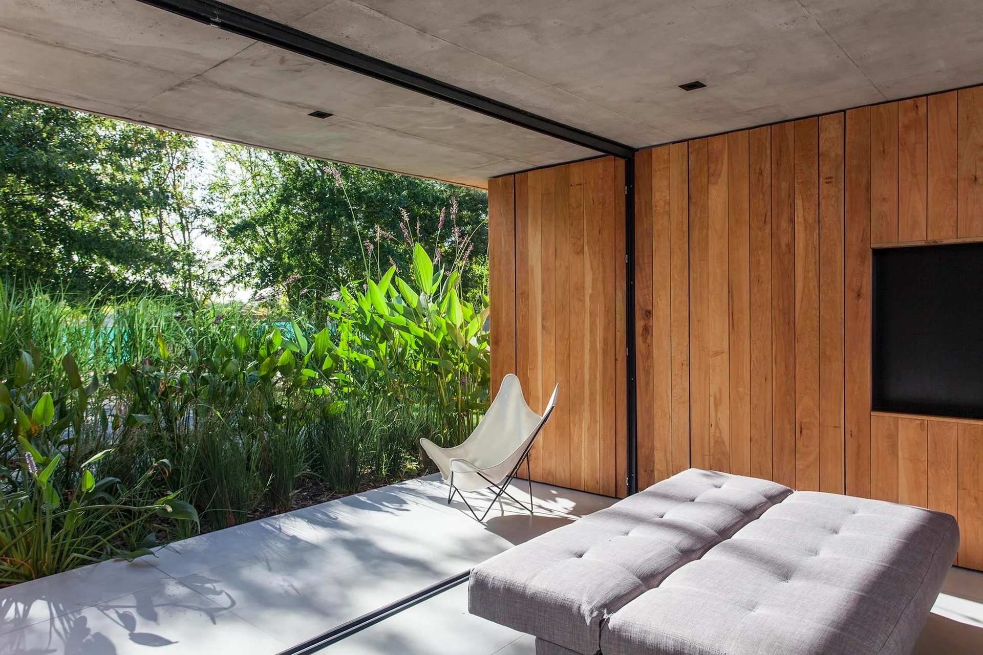A modern indoor-outdoor patio featuring a grey tufted daybed and white butterfly chair set against vertical wood paneling and a concrete ceiling.