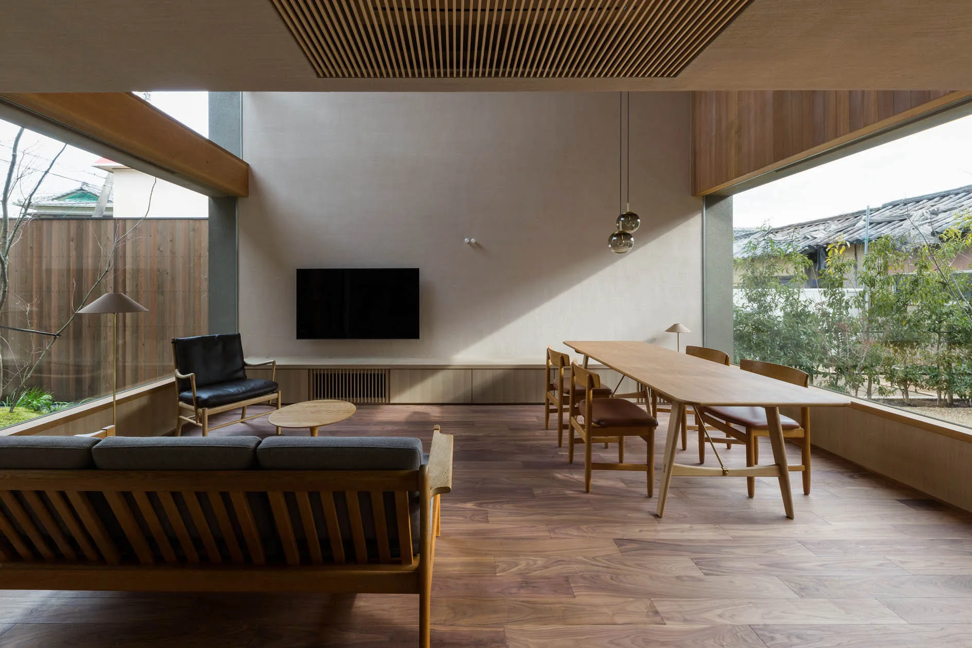 A spacious living and dining room features a long wooden dining table, a low-profile sofa, a wall-mounted TV, and large windows looking outside.