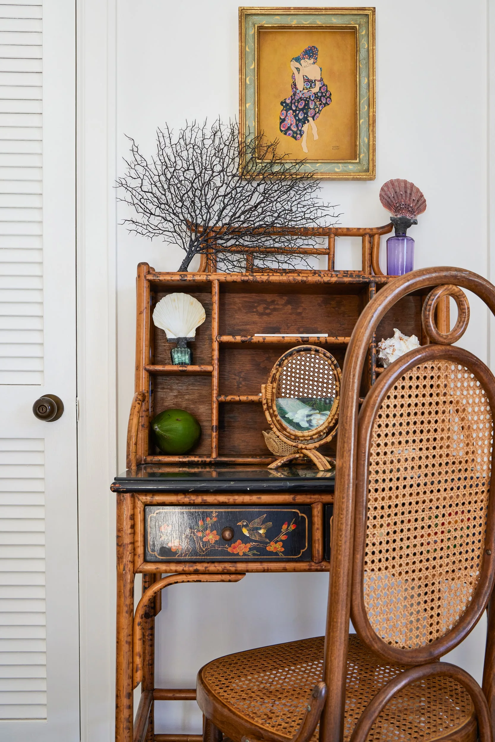 A vintage bamboo writing desk with small shelves and a cane chair sit next to a white louvered door.