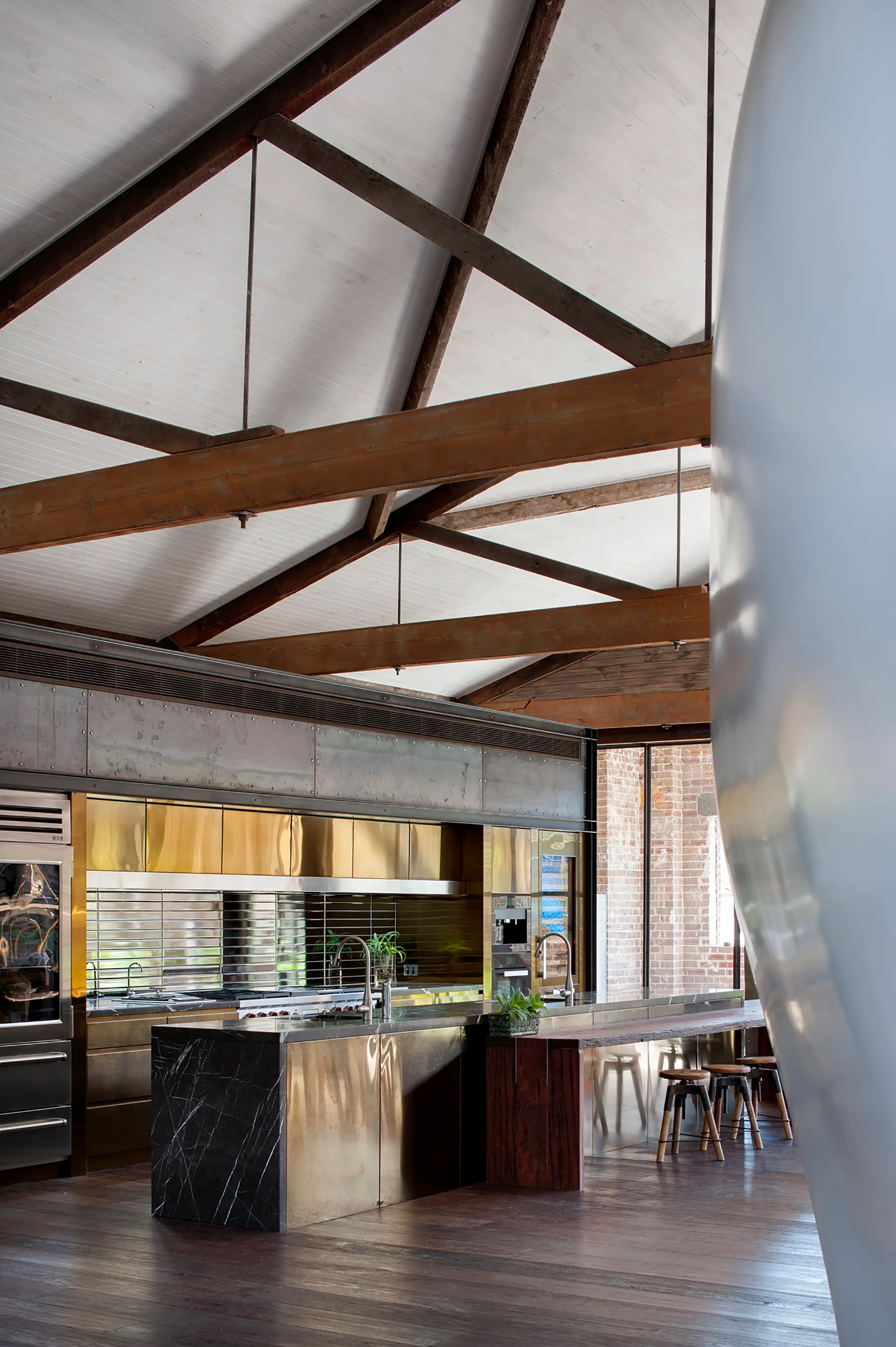 A spacious kitchen featuring polished brass cabinets, a stone island, and a vaulted ceiling with exposed timber beams over dark wood floors.