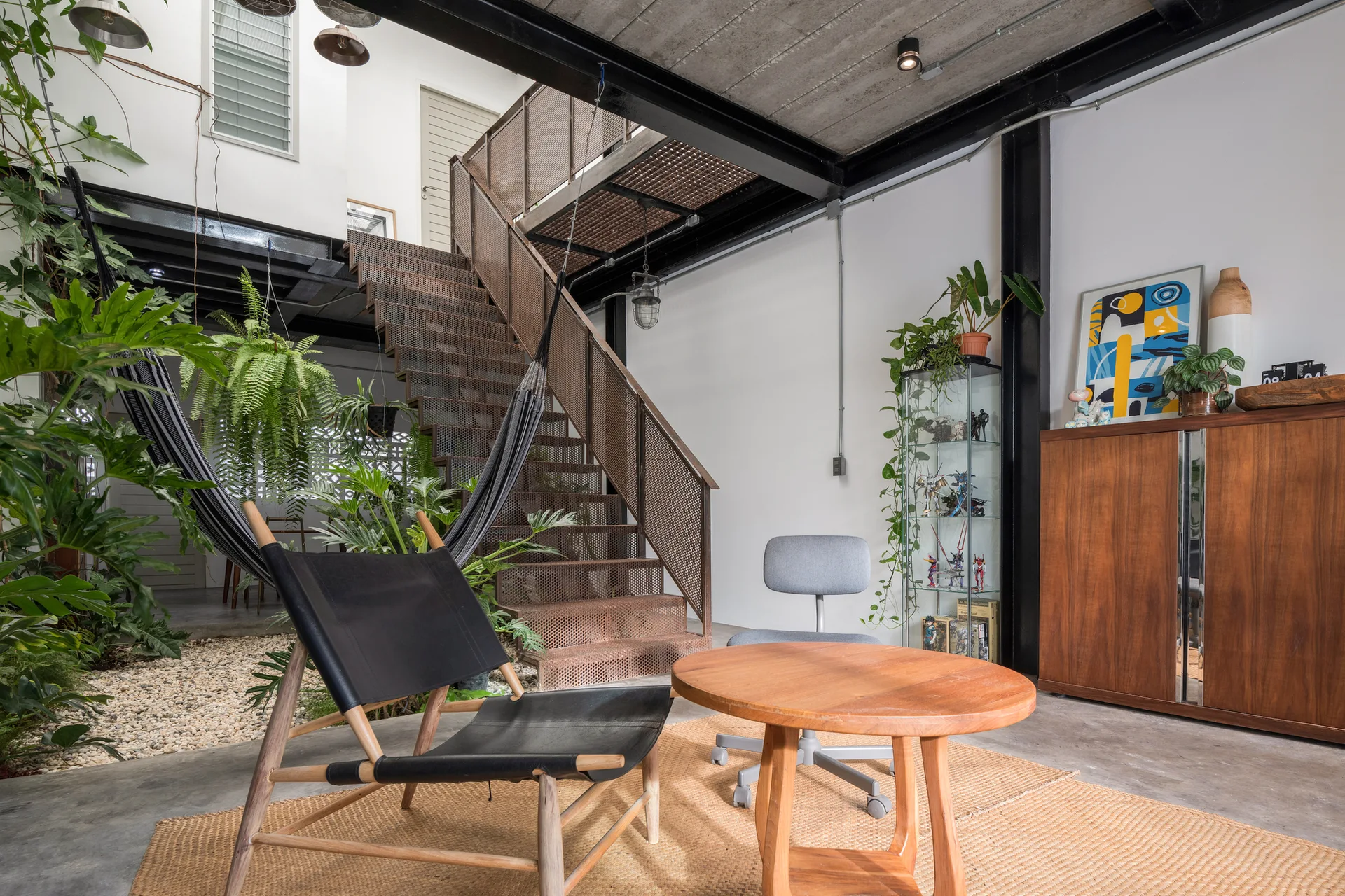 An industrial living room features a perforated metal staircase, a gravel garden bed with plants, a black hammock, and wood furniture.