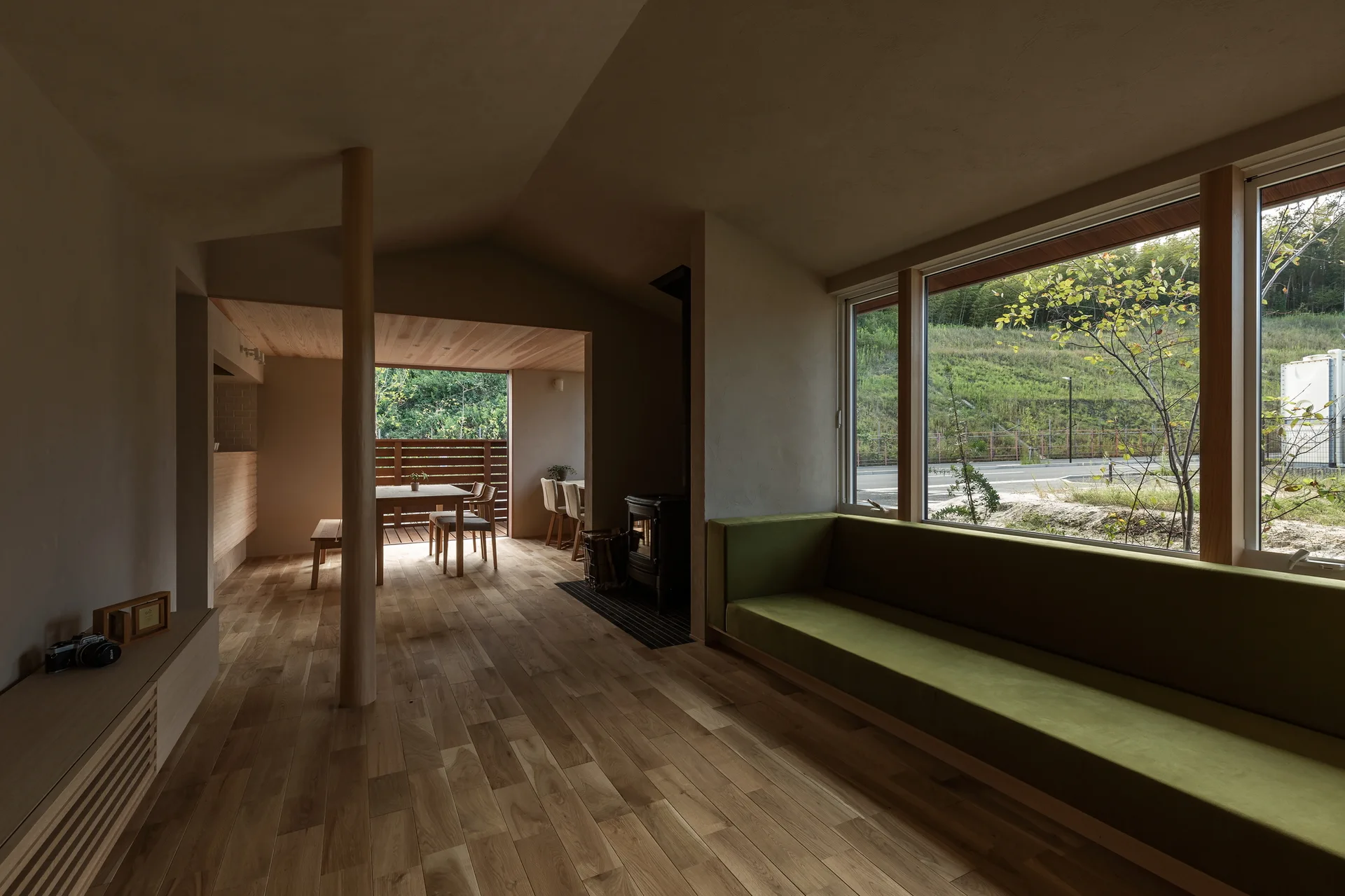 A minimalist open-plan living room features a long green built-in sofa below a large window, natural wood floors, and a dining table in the background.