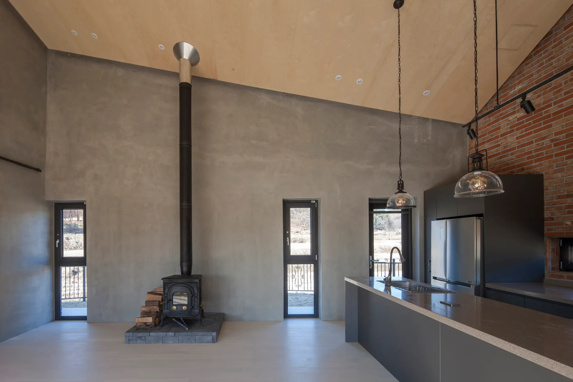 An industrial kitchen features a concrete wall with a black wood stove, a dark grey island, and a sloped plywood ceiling.