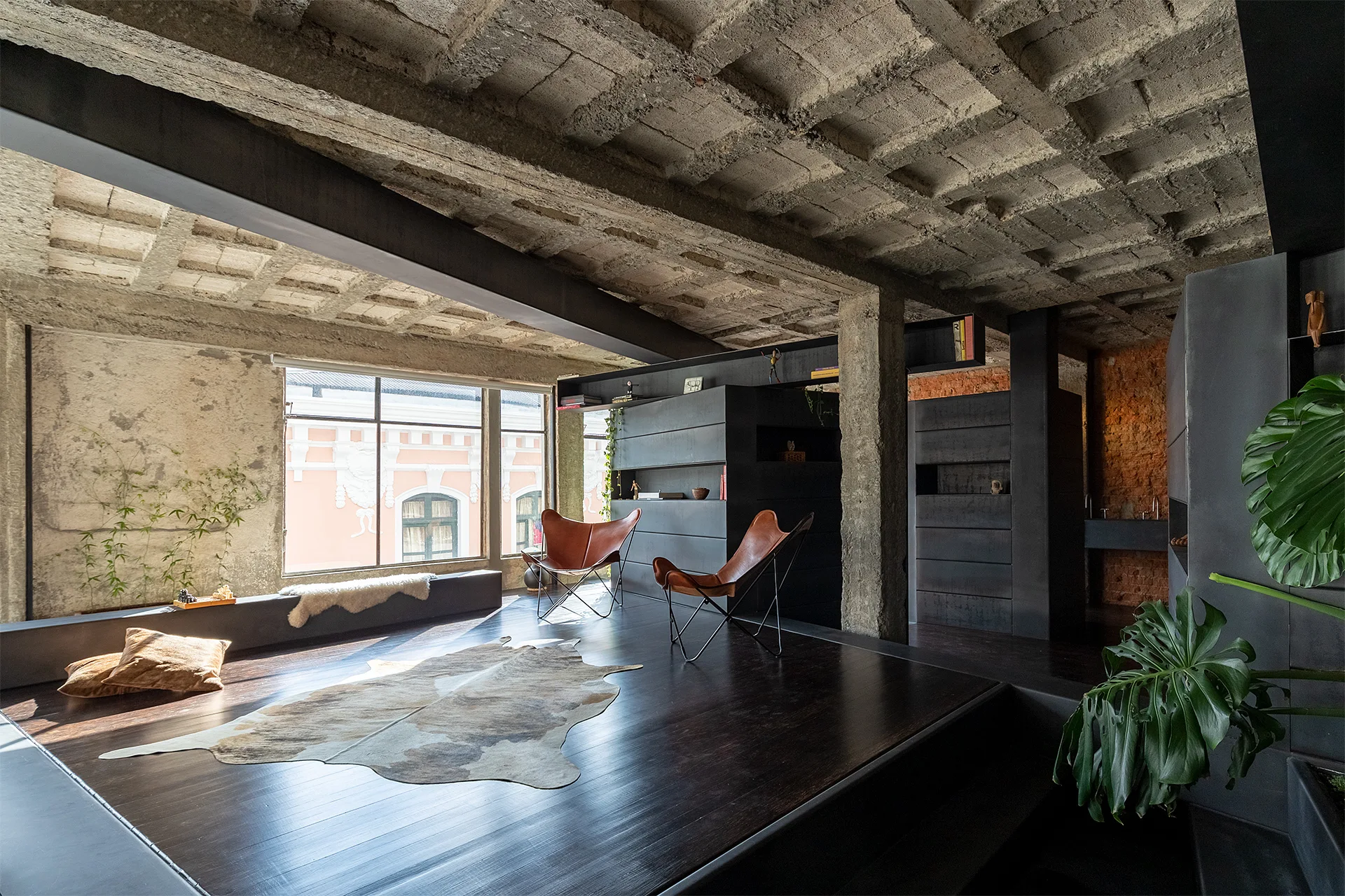 An industrial loft living room featuring an exposed concrete waffle ceiling, dark wood floors, a cowhide rug, and two tan leather butterfly chairs.