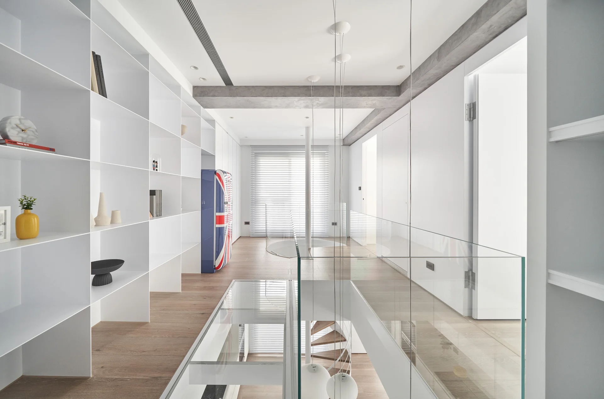 A minimalist white hallway features floor-to-ceiling built-in shelving, wood flooring with a glass insert, and a glass-paneled railing.