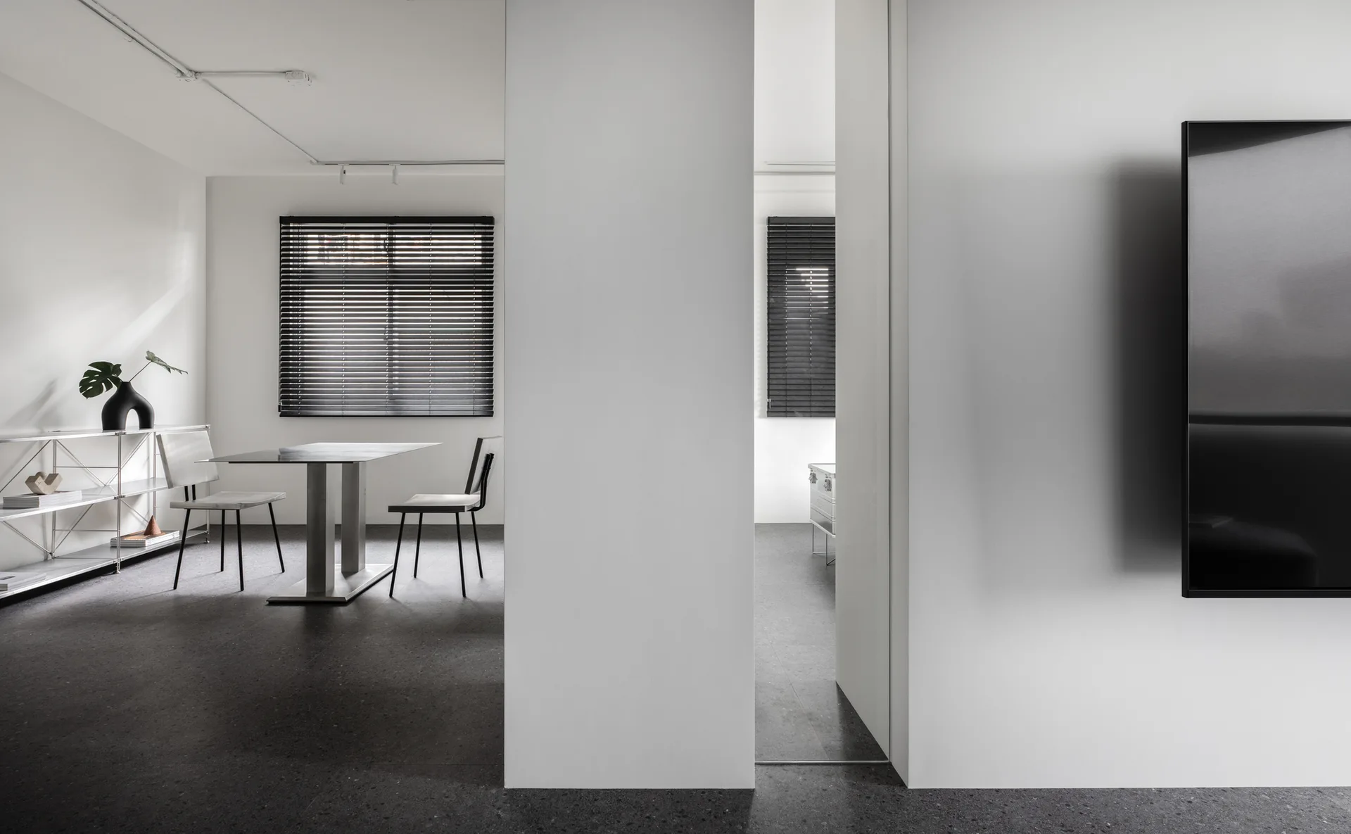 A minimalist dining room features a metal table and chairs, a wire shelving unit, and a window with dark venetian blinds set against stark white walls.