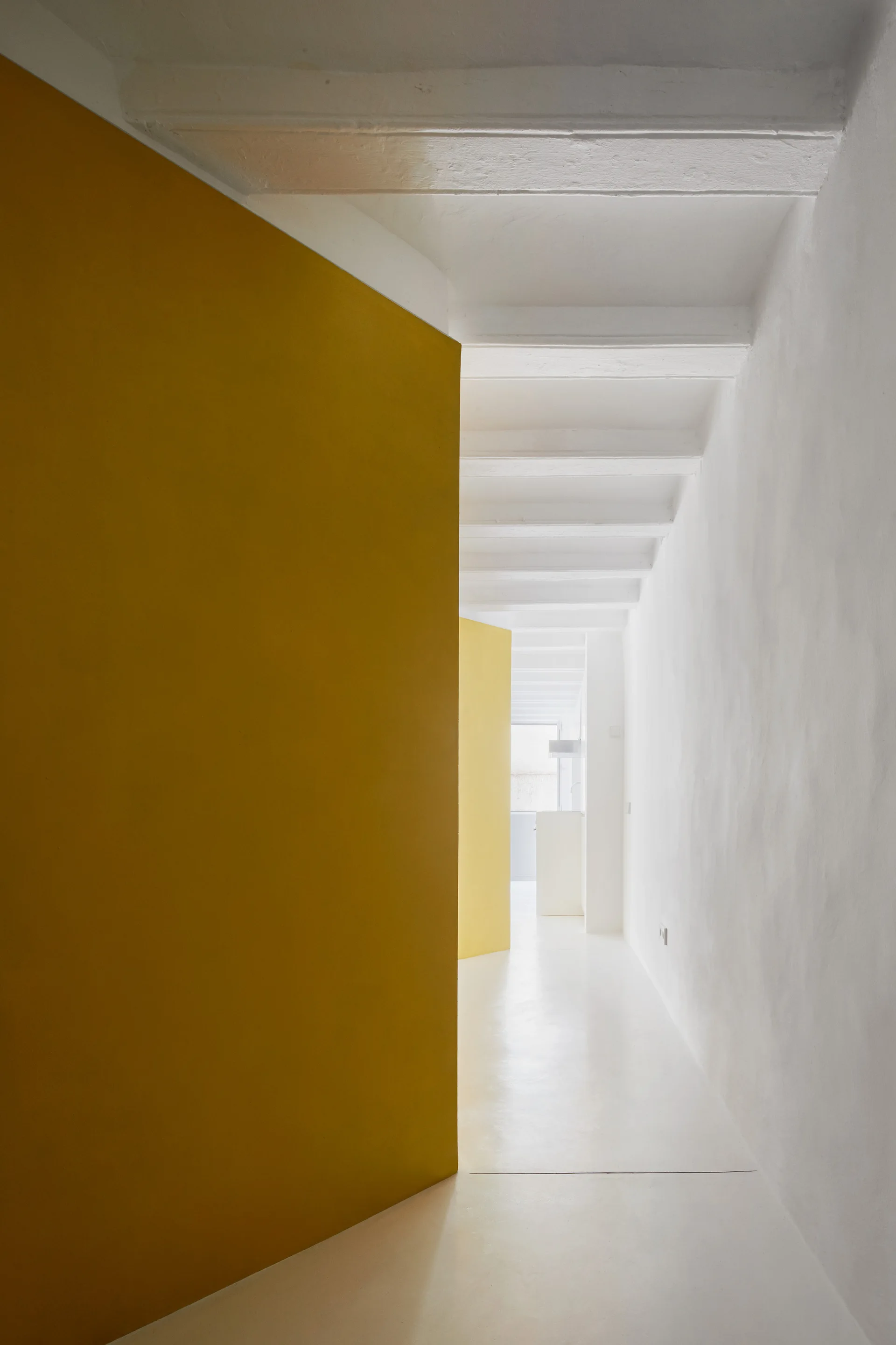 A long minimalist white hallway features a prominent mustard yellow partition wall, white floors, and a painted beamed ceiling.