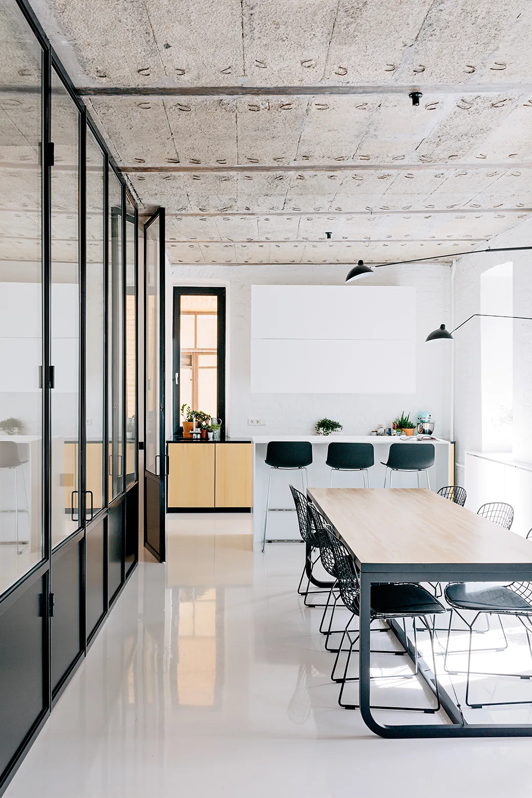 A modern dining room with a light wood table, black wire chairs, an exposed concrete ceiling, and a black-framed glass partition wall.