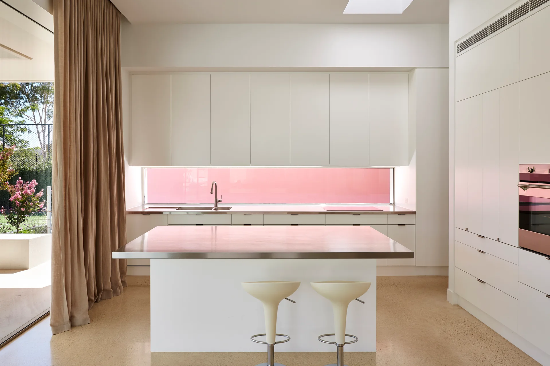 A minimalist white kitchen features a stainless steel island countertop and a long horizontal window looking out to a bright pink wall.