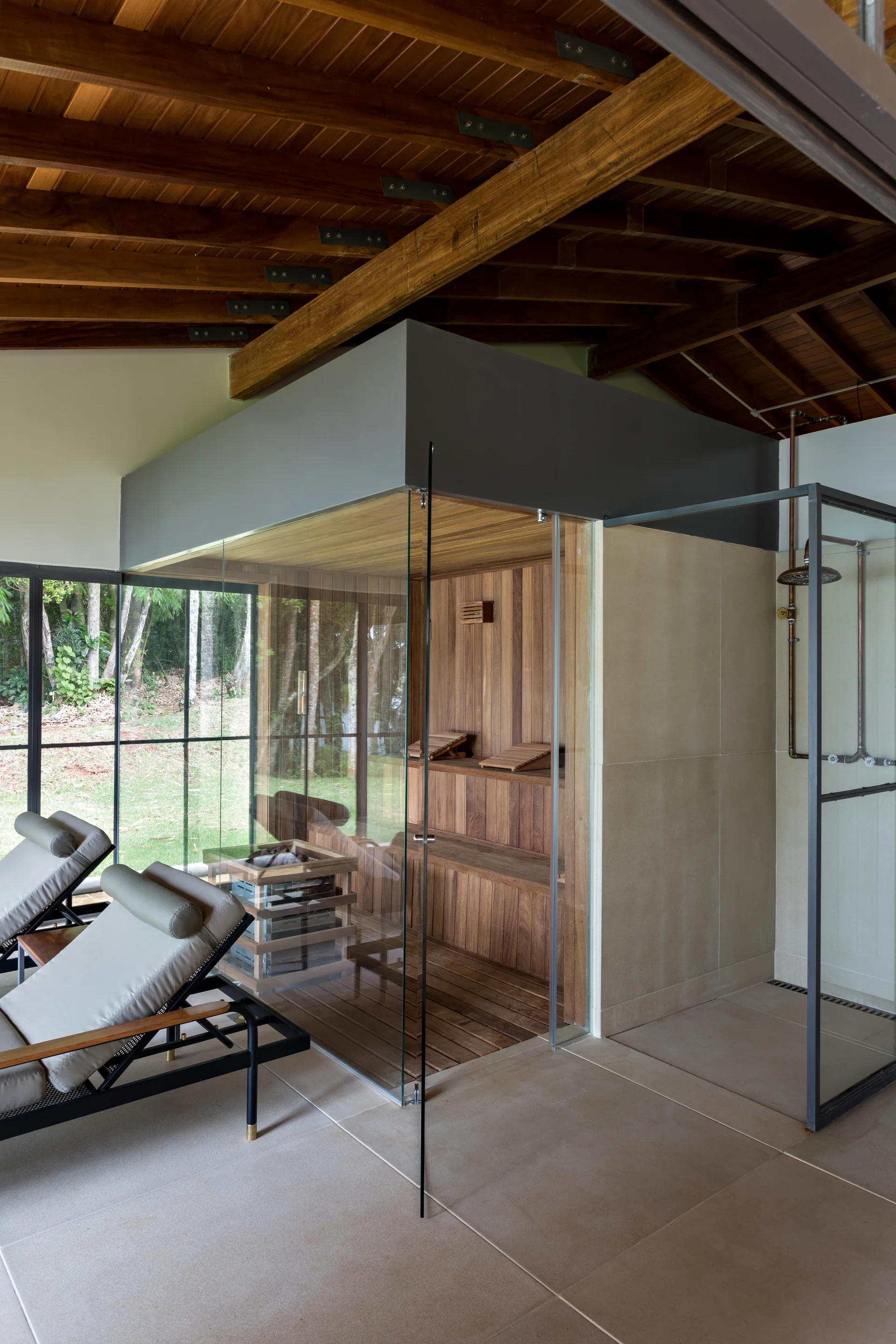 An indoor wellness room features a glass-enclosed wood sauna, a tiled shower with copper fixtures, and two lounge chairs beneath a beamed timber ceiling.