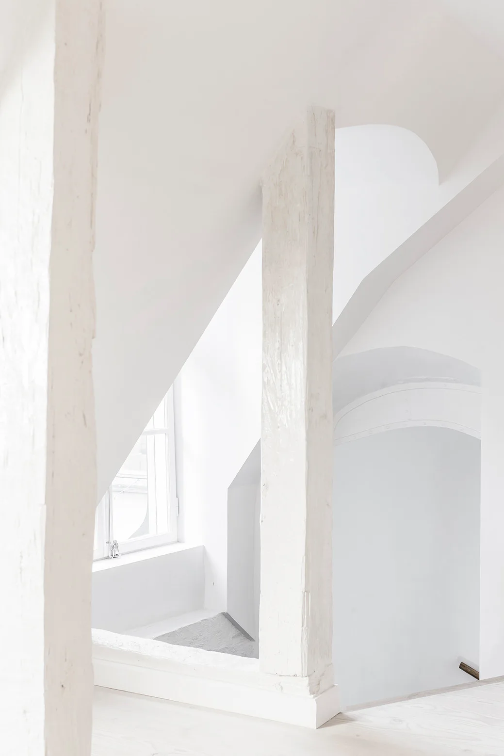 A bright, all-white minimalist stairwell featuring painted vertical wooden beams, geometric angled walls, and a small window letting in natural light.