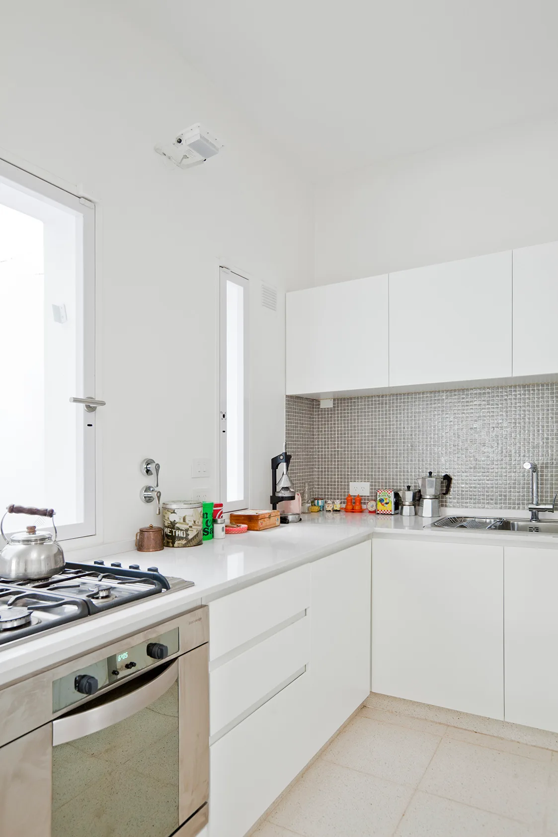 A minimalist white kitchen features flat cabinetry, a grey mosaic tile backsplash, two slim vertical windows, and stainless steel appliances.